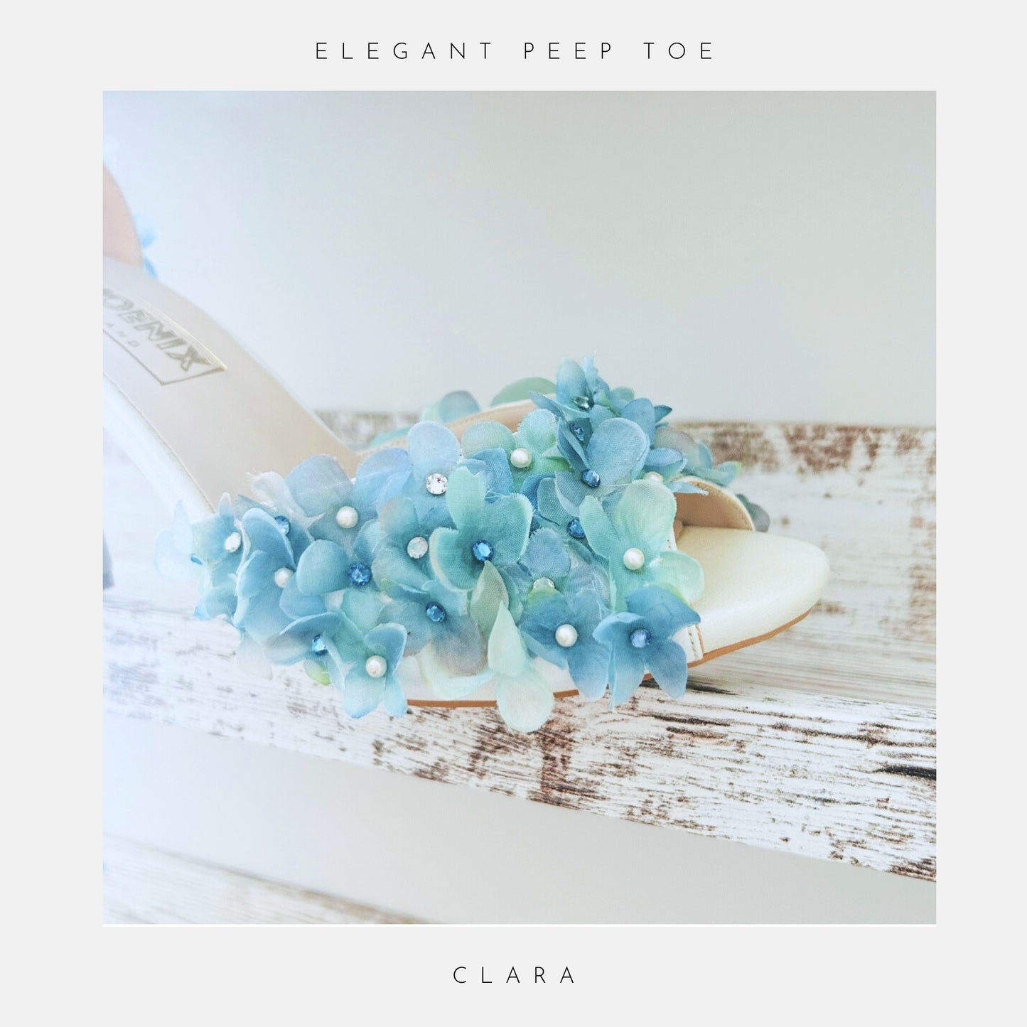 Ivory wedding sandal with blue floral decorations on a wooden surface, labelled 'Elegant Peep Toe Clara'.