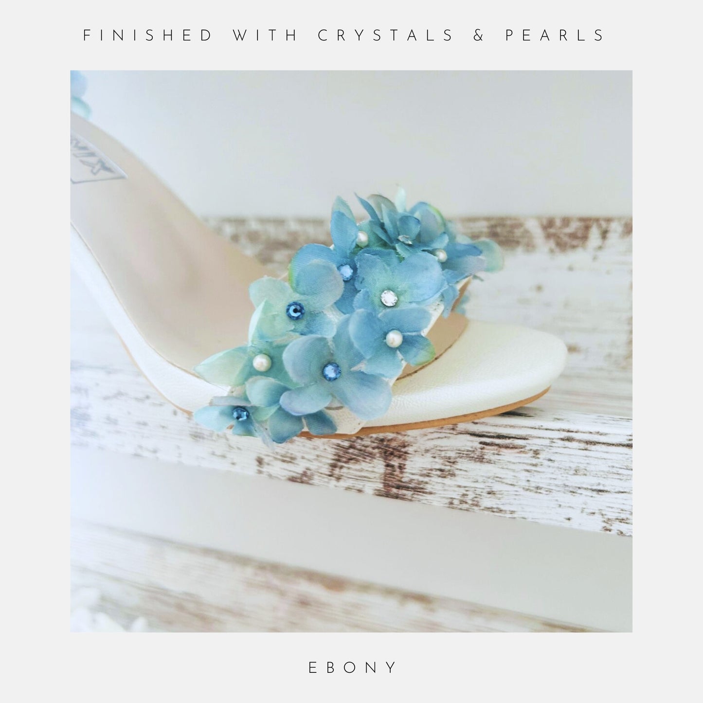 Ivory sandal with blue floral embellishments on a wooden surface, labelled 'Finished With Crystals & Pearls' and 'Ebony'.