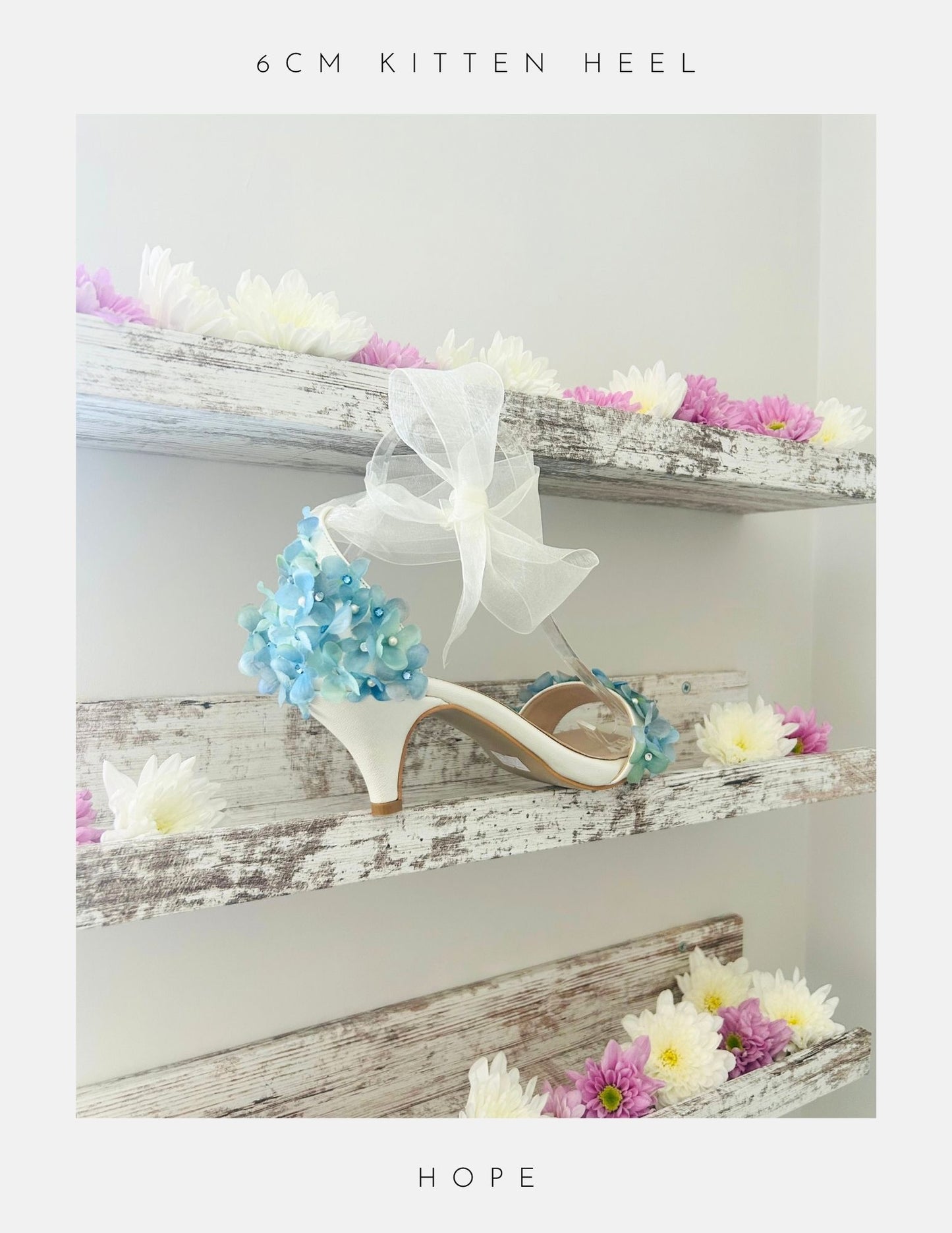 The Hope Organza Floral Kitten Heel Bridal Shoes by Phoenix England feature white kitten heels, blue organza floral accents, and a large white bow, displayed on rustic shelves with pink and white flowers. Text above: 6CM KITTEN HEEL. Below: HOPE.