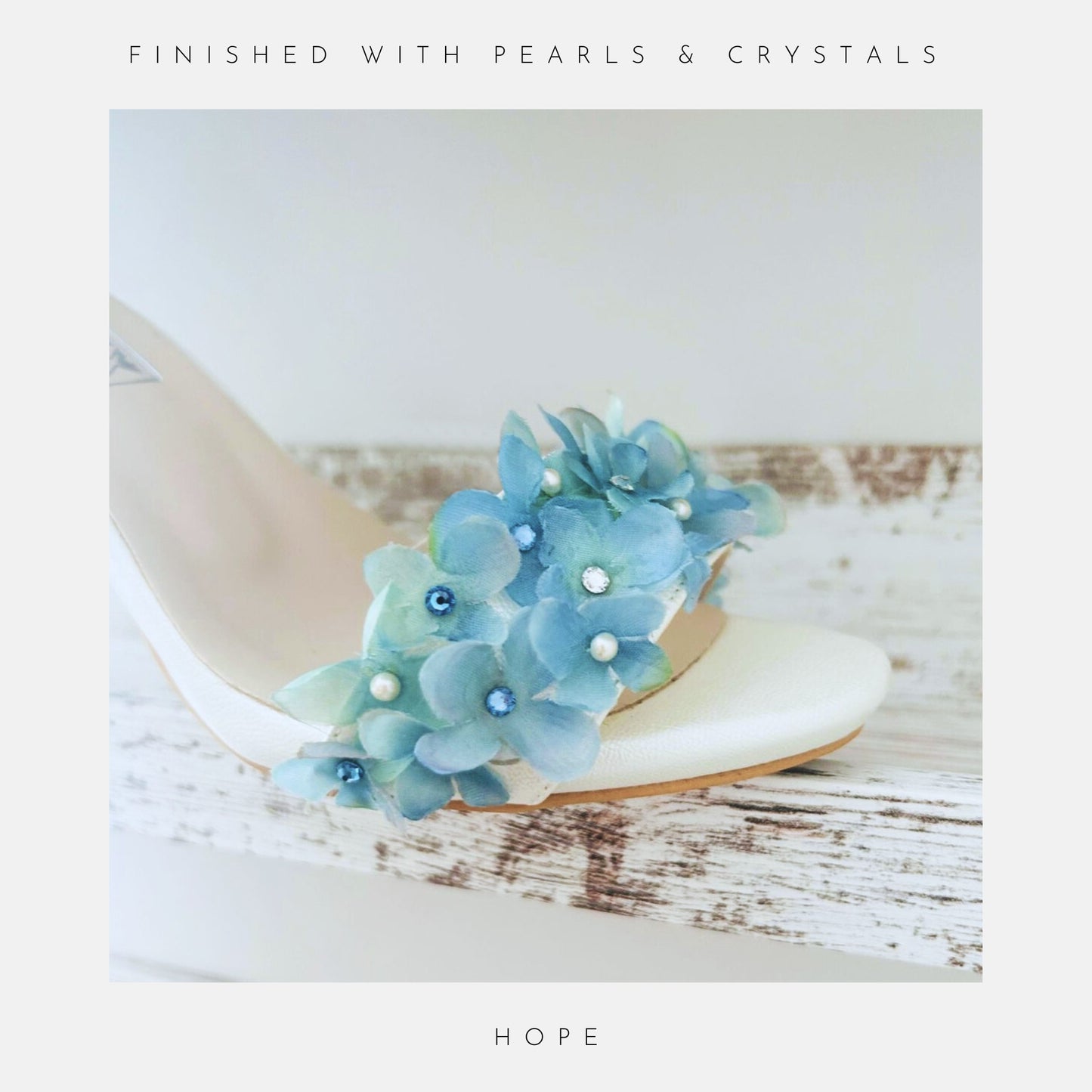 A close-up of Phoenix England’s Hope Organza Floral Kitten Heel Bridal Shoes, featuring blue organza flowers, Austrian crystals & pearls, on a rustic white wood surface. AUSTRIAN CRYSTALS & PEARLS tops the image; HOPE appears below.