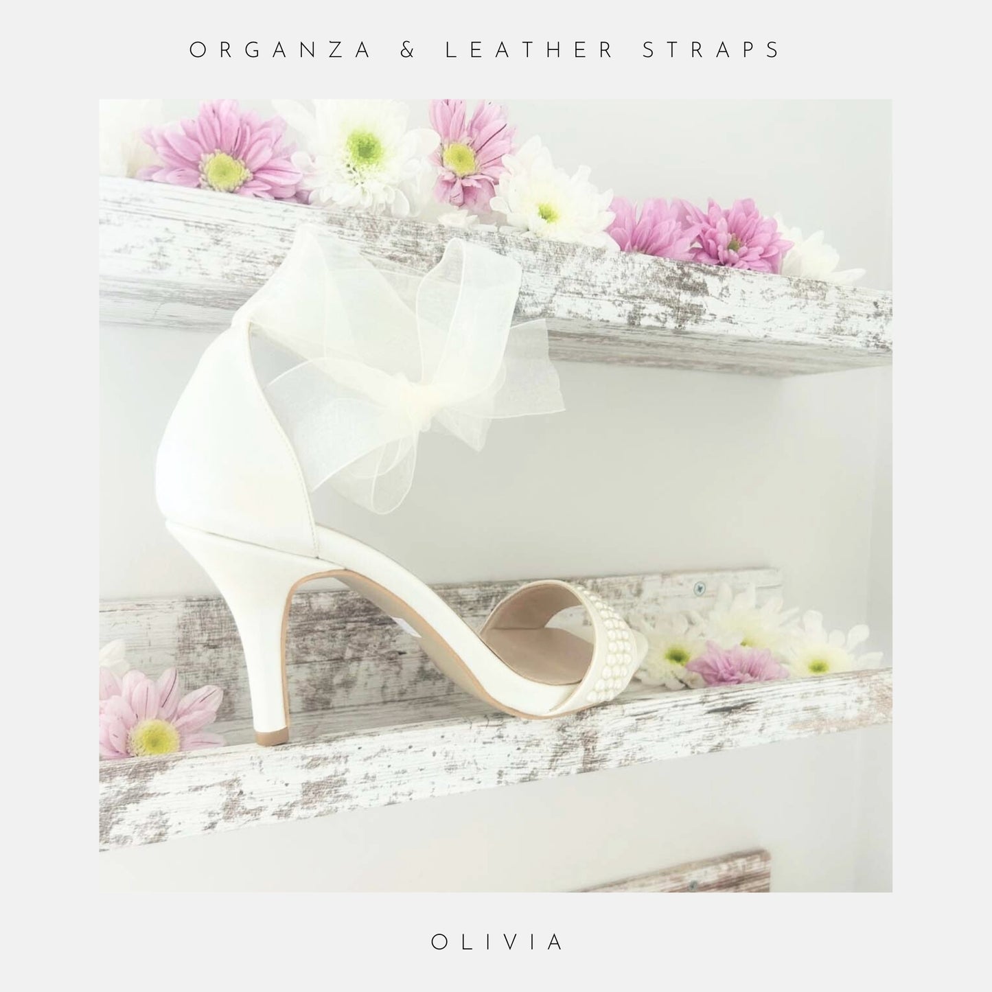 An ivory Olivia Organza Strap Pearl Bridal Shoe by Phoenix England rests on a rustic white wooden shelf with pink and white flowers. Above: “organza and leather straps.” Below: “OLIVIA.”.