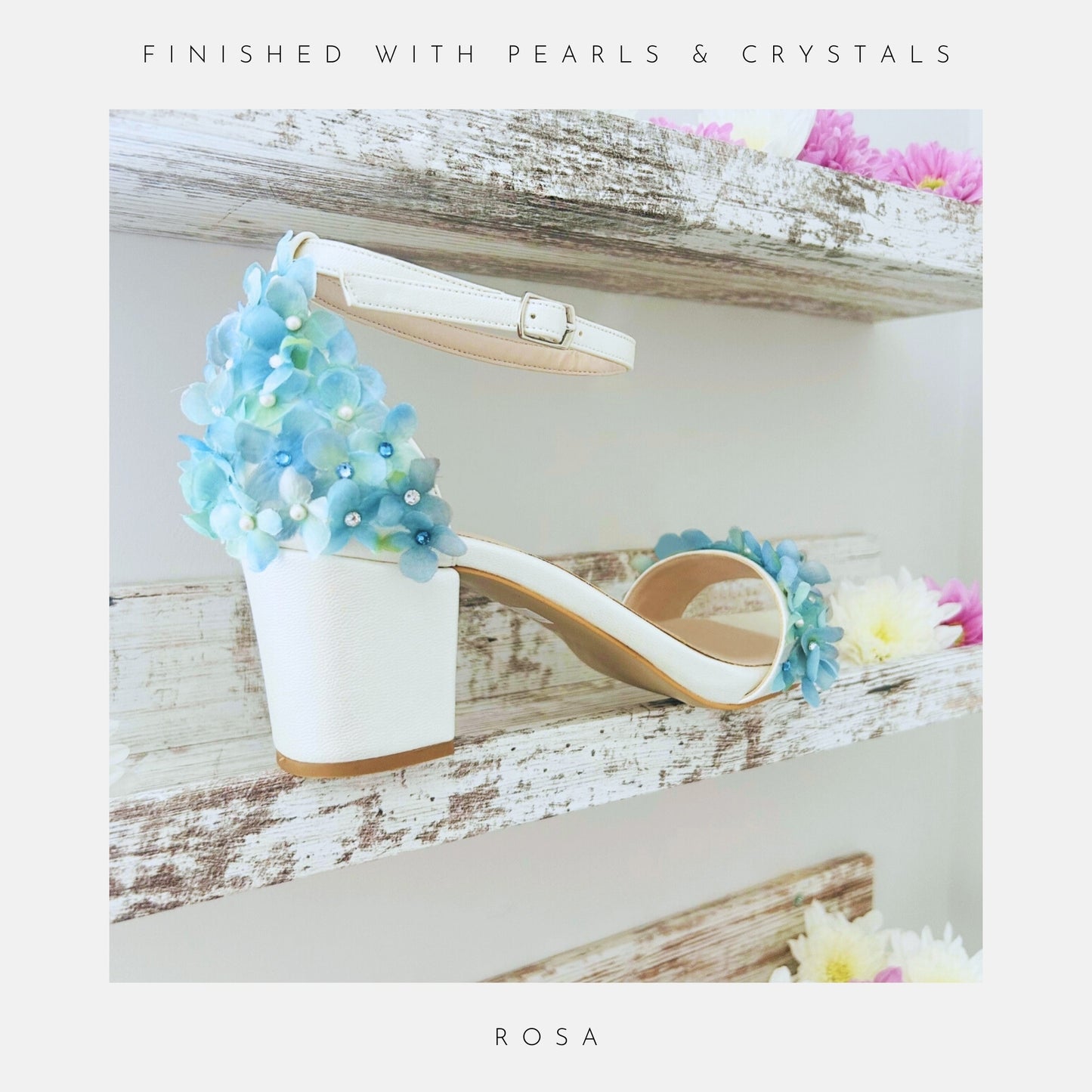 A white 6CM Rosa Floral Low Block Heel Bridal Shoe by Phoenix England with blue and white floral heel embellishments sits on a rustic shelf decorated with colourful flowers, perfect chic bridal shoes.