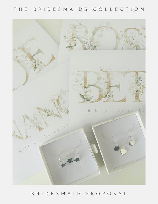 Flat lay of Phoenix England Be My Bridesmaid Gifts: proposal cards with floral lettering, next to two white sterling silver jewellery boxes—one with three star charms, the other with two heart charms.