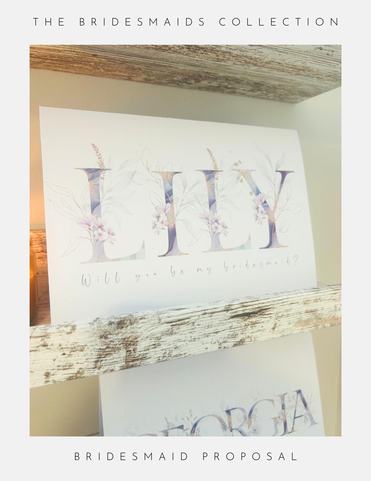 A Phoenix England Bridesmaids Proposal Sterling Silver card with soft florals and “Will you be my bridesmaid?” for Lily sits on a rustic wooden shelf, while another card peeks below with partial name and bordered text.