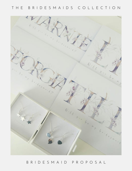 A flat lay of Phoenix England Bridesmaids Proposal Sterling Silver necklaces—stars, hearts, and circles—in white boxes with personalized floral Will you be my bridesmaid? cards. Text: The Bridesmaids Collection.