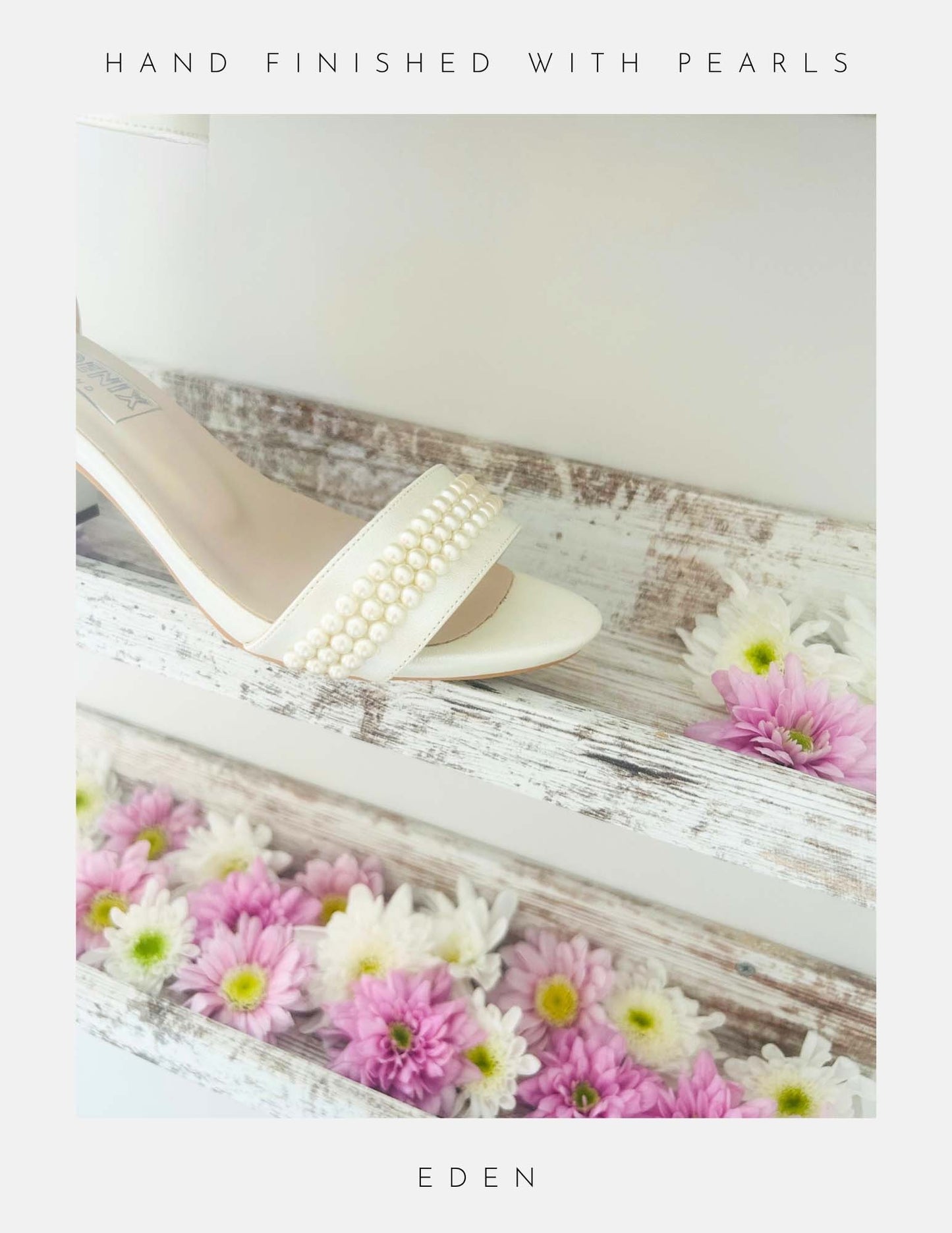 A pair of Eden Pearl Low Block Heel Bridal Shoes by Phoenix England, in ivory with pearl embellishments, rests on a rustic white wooden shelf adorned with pink and white daisies. Text border: HAND FINISHED WITH PEARLS and EDEN.