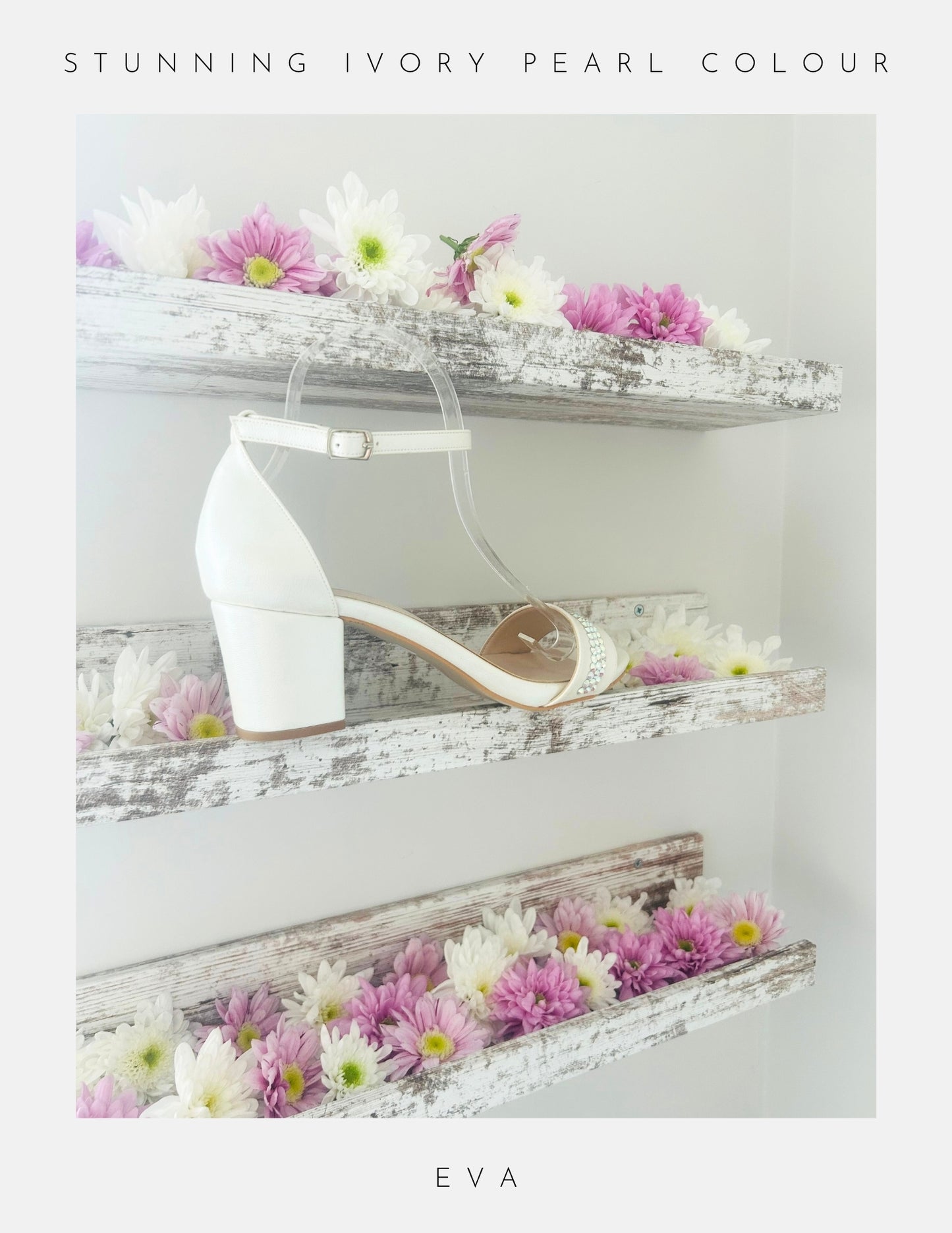 The Eva Crystal Low Block Heel Bridal Shoes by Phoenix England are ivory pearl vegan leather sandals with ankle straps and low block heels, displayed on shabby-chic wooden shelves adorned with white and pink flowers.