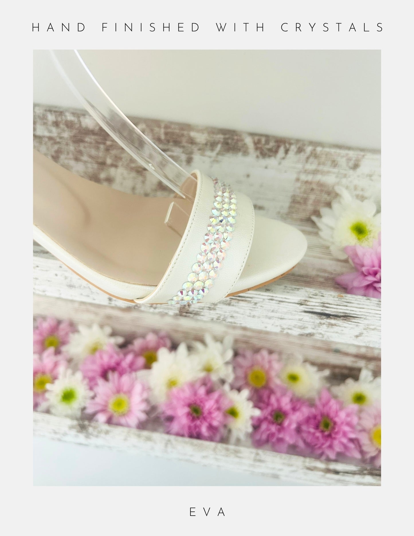 A close-up of the Eva Crystal Low Block Heel Bridal Shoes by Phoenix England, crafted from vegan leather with sparkling crystals on the strap, shown on whitewashed wood with pink and white daisies. Text above: Hand finished with crystals; below: Phoenix England.