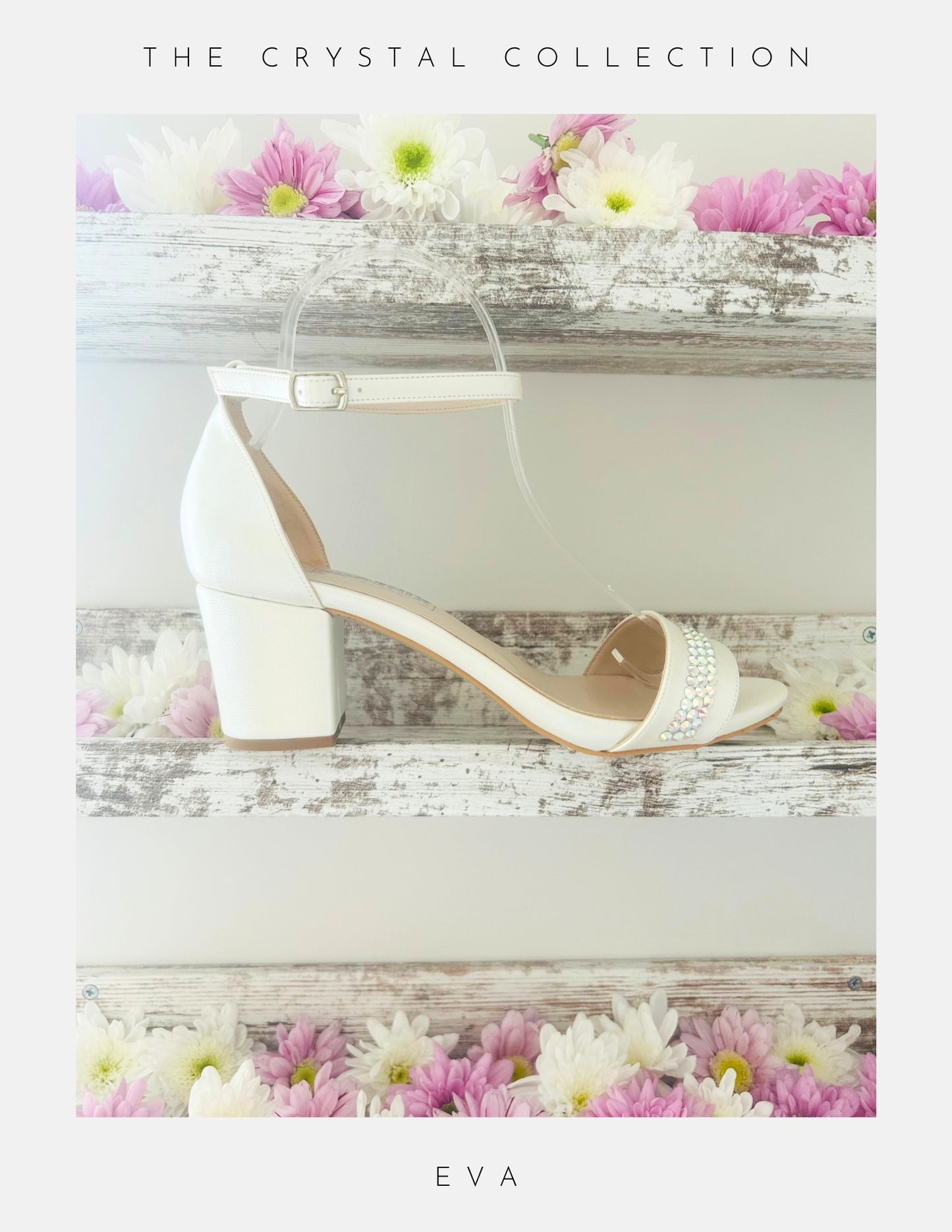 Displayed on a rustic white wooden shelf with pink and white flowers, the Phoenix England Eva Crystal Low Block Heel Bridal Shoes in vegan leather feature a white ankle strap and crystal accents. Text above: The Crystal Collection; below: Eva.