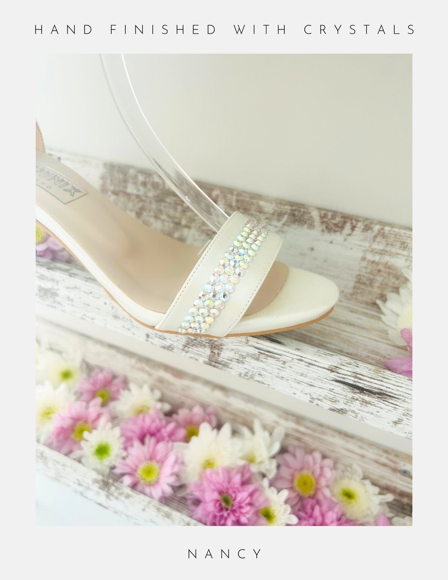 A cream sandal, the Nancy Crystal Kitten Heel Bridal Shoes by Phoenix England, features pearl vegan leather and a crystal-embellished strap. Displayed on a rustic white shelf with pastel daisies. “HAND FINISHED WITH CRYSTALS” above and “NANCY” below. Perfect for weddings.