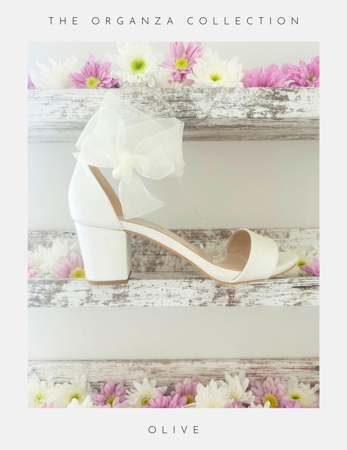 The Phoenix England Olive Block Heel Bridal Shoes Organza Strap, an ivory block heel bridal shoe with an organza bow, is displayed on a rustic white wooden shelf with pink and white flowers. Text: The Organza Collection.