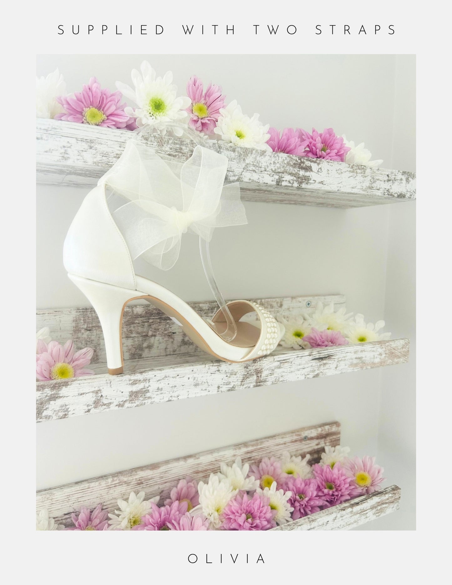 An ivory Olivia Organza Strap Pearl Bridal Shoe by Phoenix England rests on a rustic white wooden shelf with pink and white flowers. Above: “SUPPLIED WITH TWO STRAPS.” Below: “OLIVIA.”.