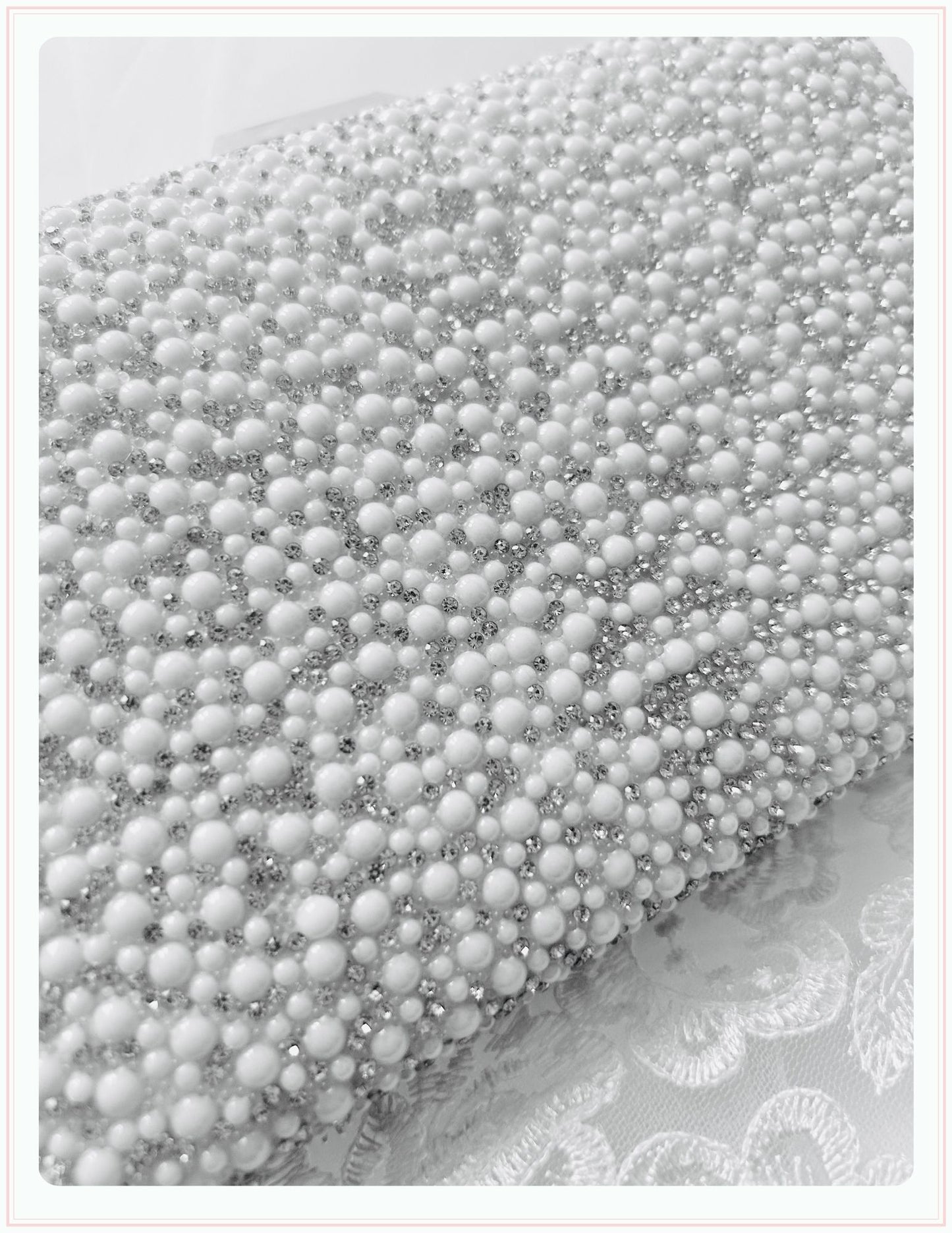 A black and white close-up of the Phoenix England Pearlita Wedding Handbag, its surface adorned with clusters of white pearls and small crystals, resting on delicate embroidered lace fabric.