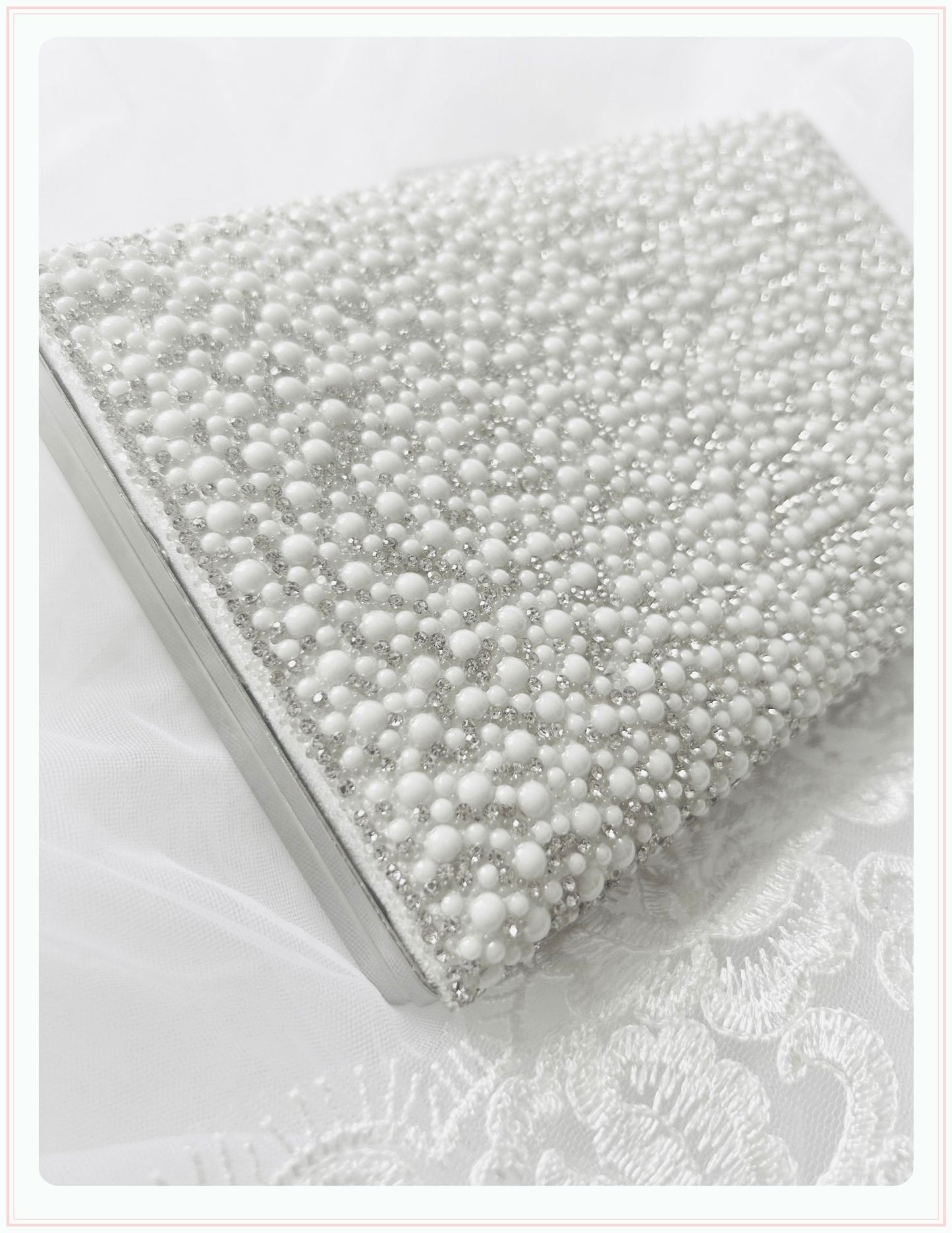 A close-up of the Phoenix England Pearlita Wedding Handbag, a rectangular clutch embellished with white and silver beads, rests on white lace—an elegant accessory perfect for special occasions.