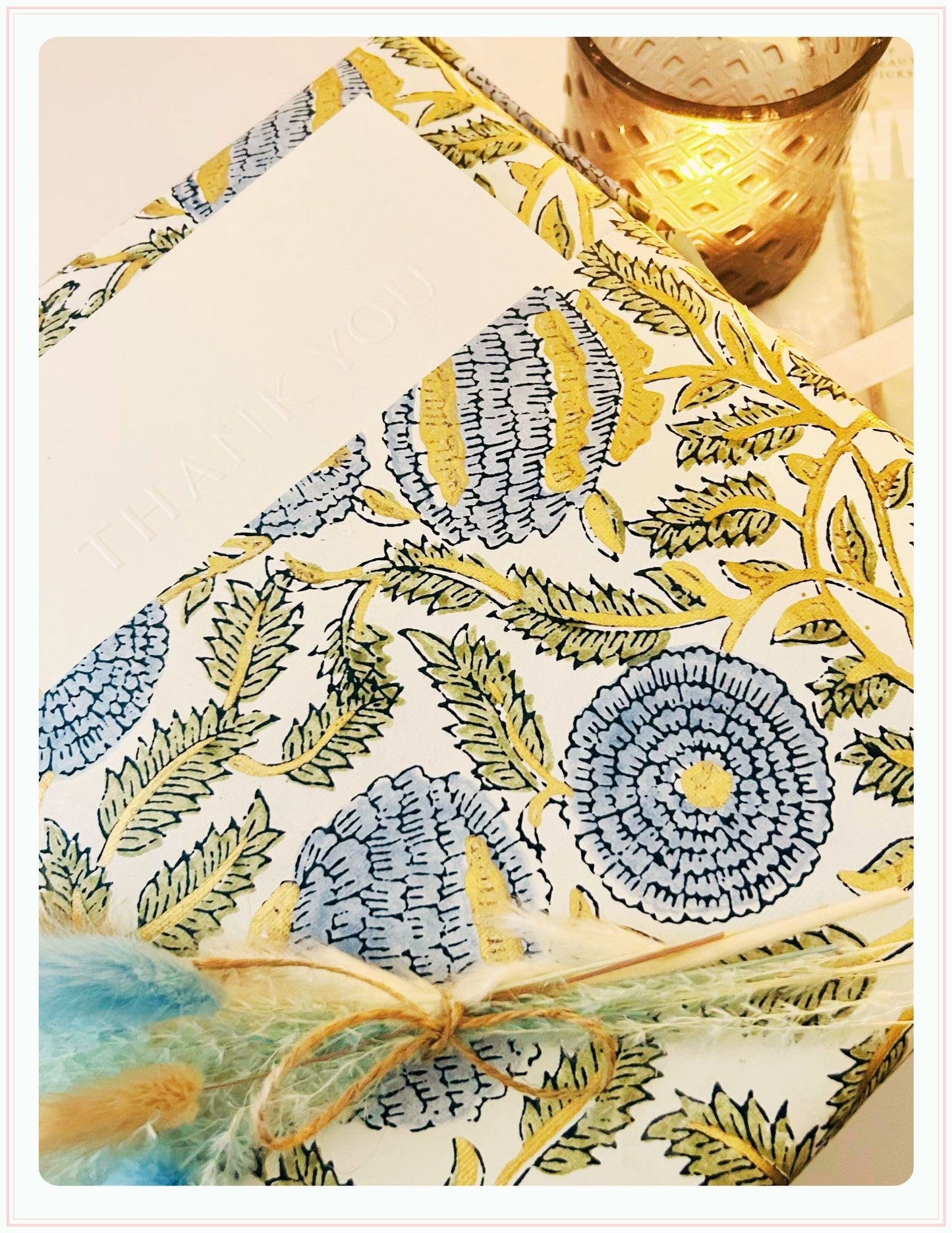 A close-up of the Phoenix England Pearlita Wedding Handbag with pearls, alongside a floral gift box labeled THANK YOU, a teal and yellow feather tied with twine, and a lit gold candle holder in the background.