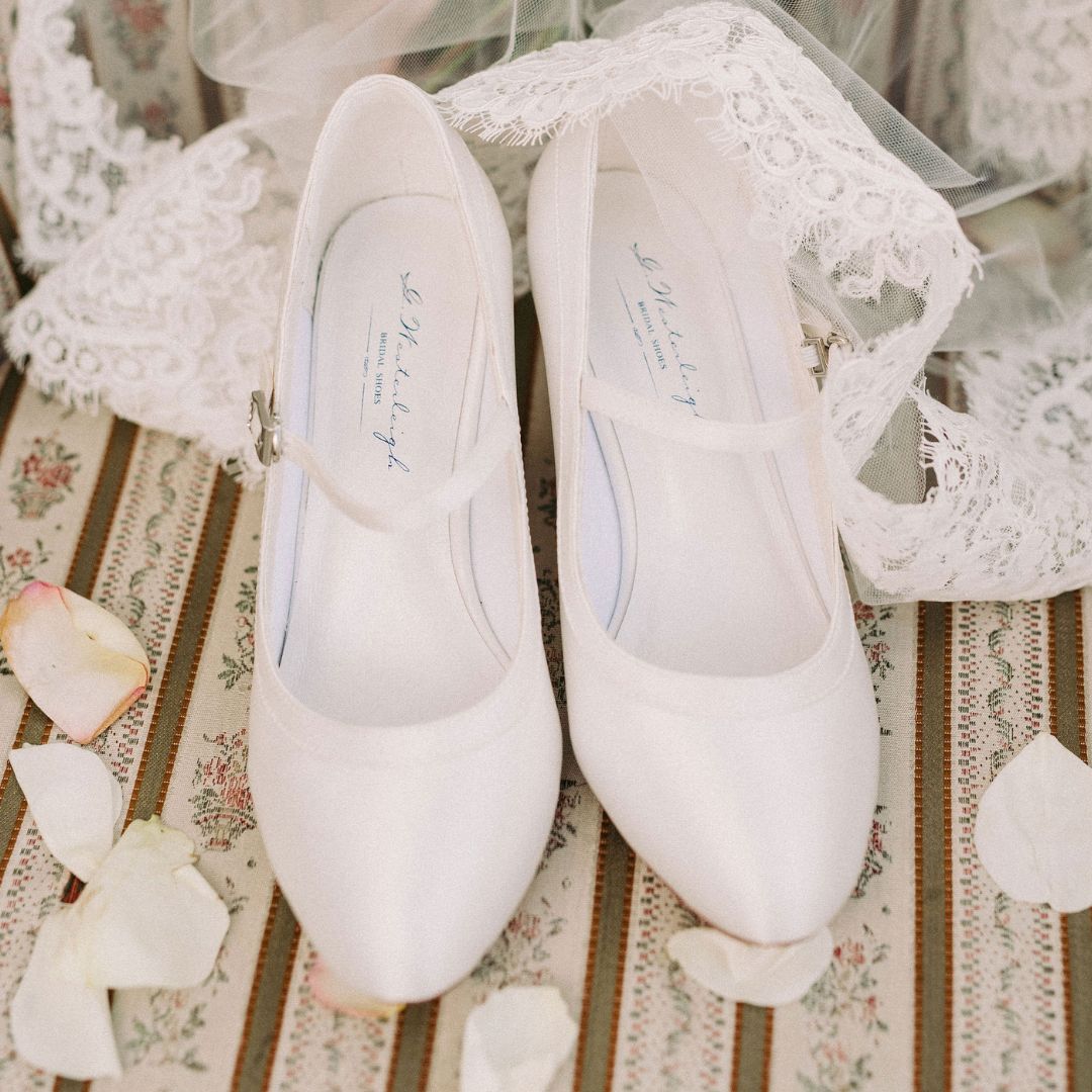Picture of a pair of ivory satin almond toe wedding shoes displayed on a chair with petals and lace.