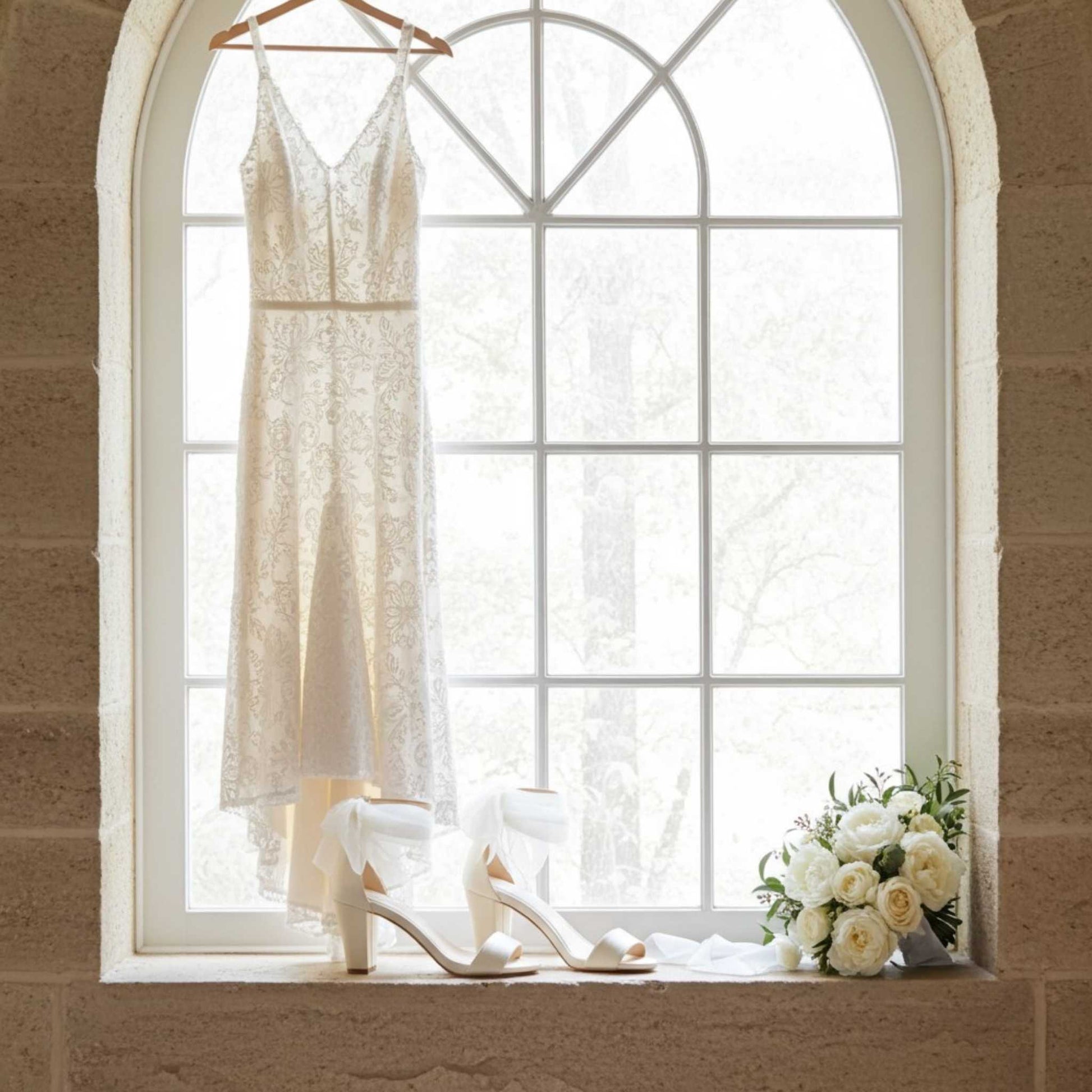 Bridal dress on a hanger with high block heel bridal shoes and flowers on a windowsill.