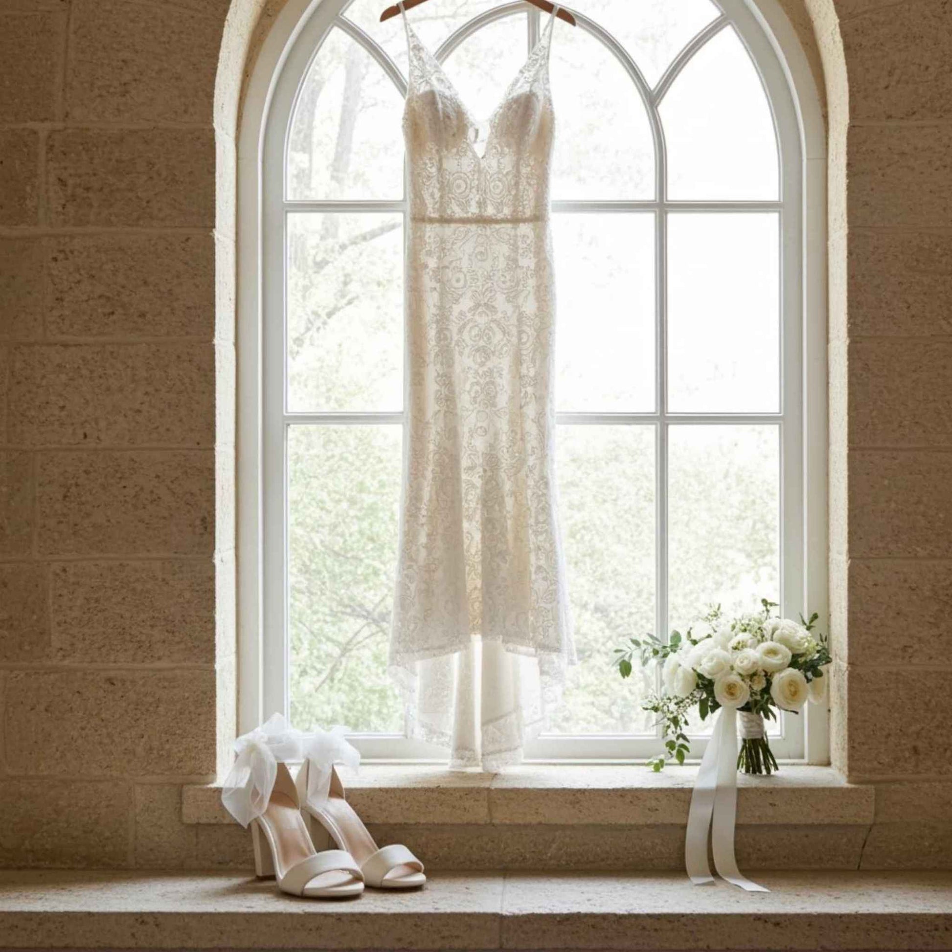 Open toe bridal shoes with organza ankle straps with bridal gown hung in window and bouquet of flowers to the side.