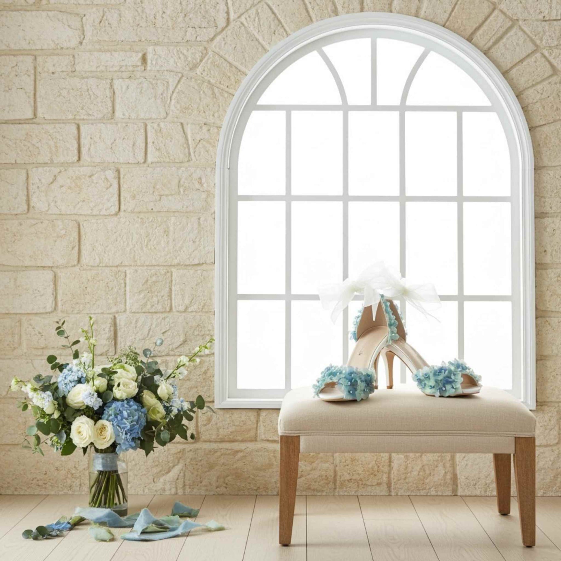 Bride's high-heeled shoe with floral decorations on a cushioned stool next to a vase of flowers against a stone wall.