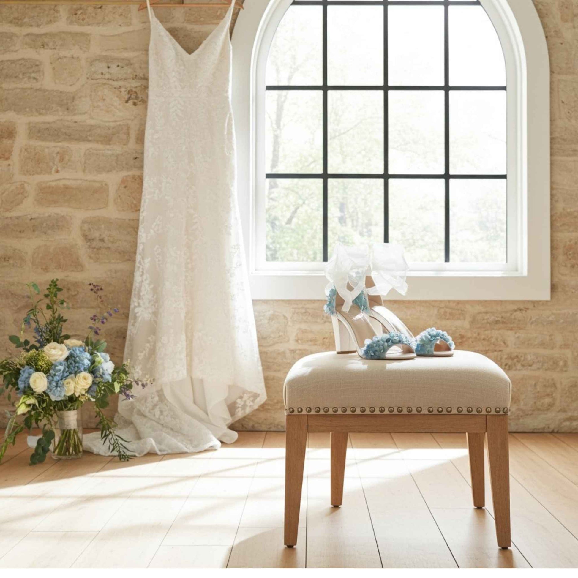 Bridal shoes decorated with flowers with a wedding gown hung behind and blue and cream floral display.