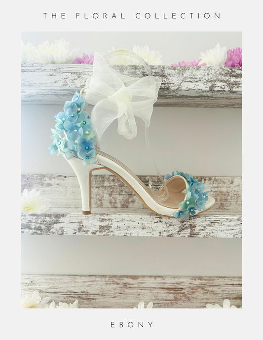 Ivory high-heeled bridal shoes with floral decorations on a wooden surface, labelled 'The Floral Collection' and 'Ebony'.