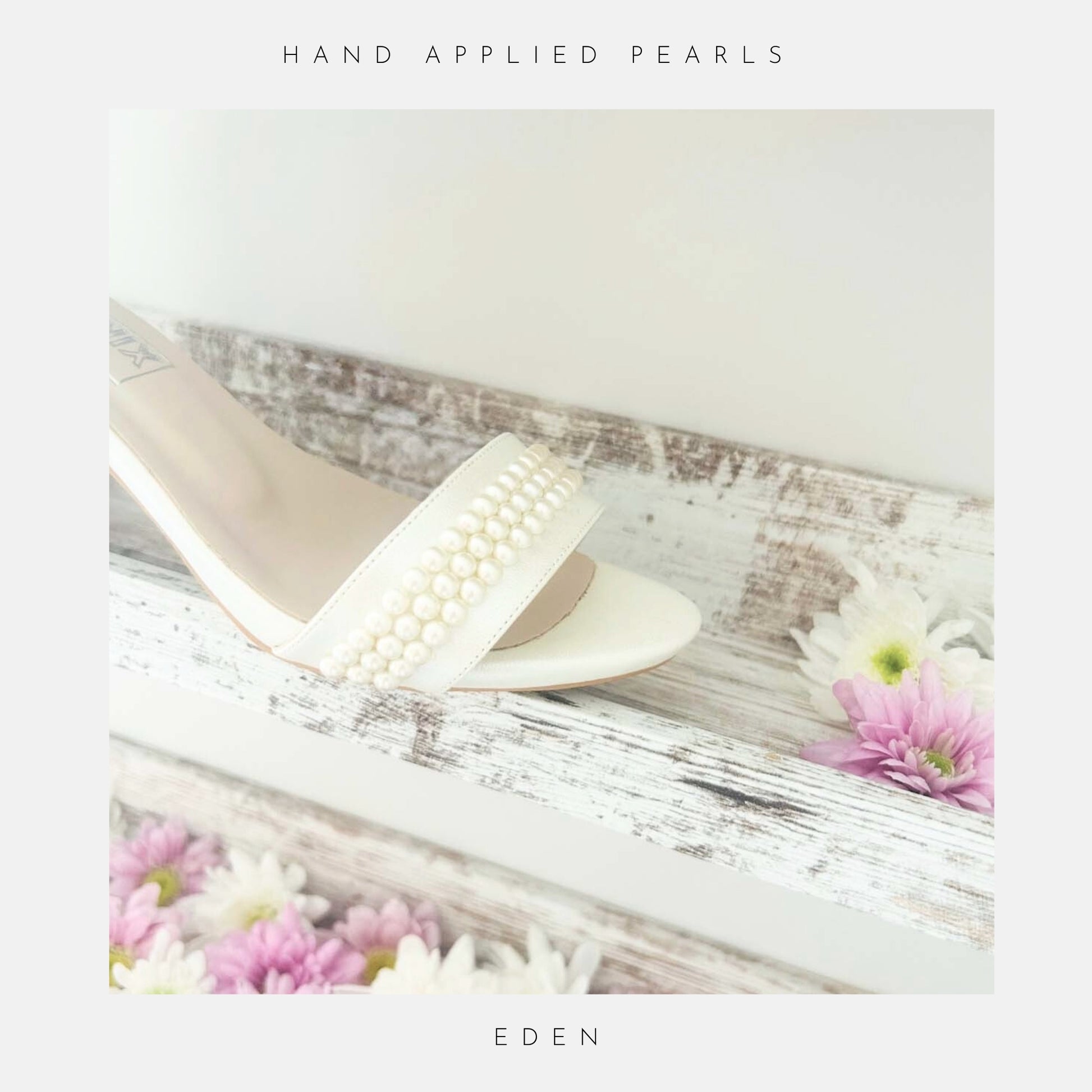 A pair of Eden Pearl Low Block Heel Bridal Shoes by Phoenix England, in ivory with pearl embellishments, rests on a rustic white wooden shelf adorned with pink and white daisies. Text border: HAND APPLIED PEARLS and EDEN.