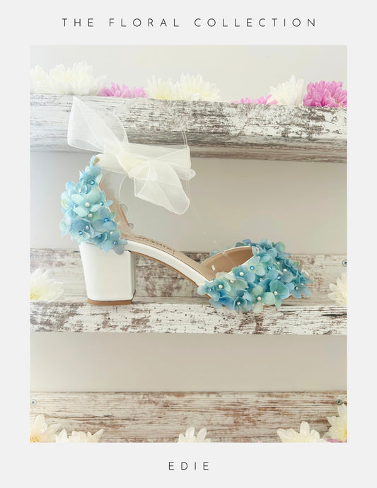 Ivory bridal sandal with blue floral decorations on a wooden surface, labelled 'The Floral Collection' and 'Edie'.