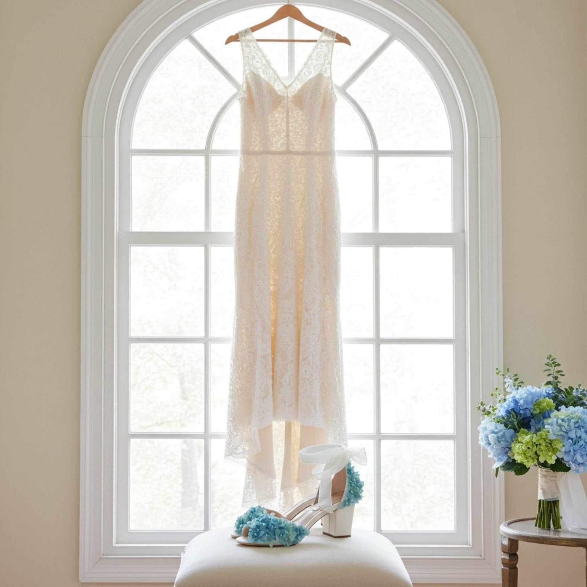 Pair of blue floral bridal shoes with low block heel and bridal gown hung in window, bouquet of hydrangeas.