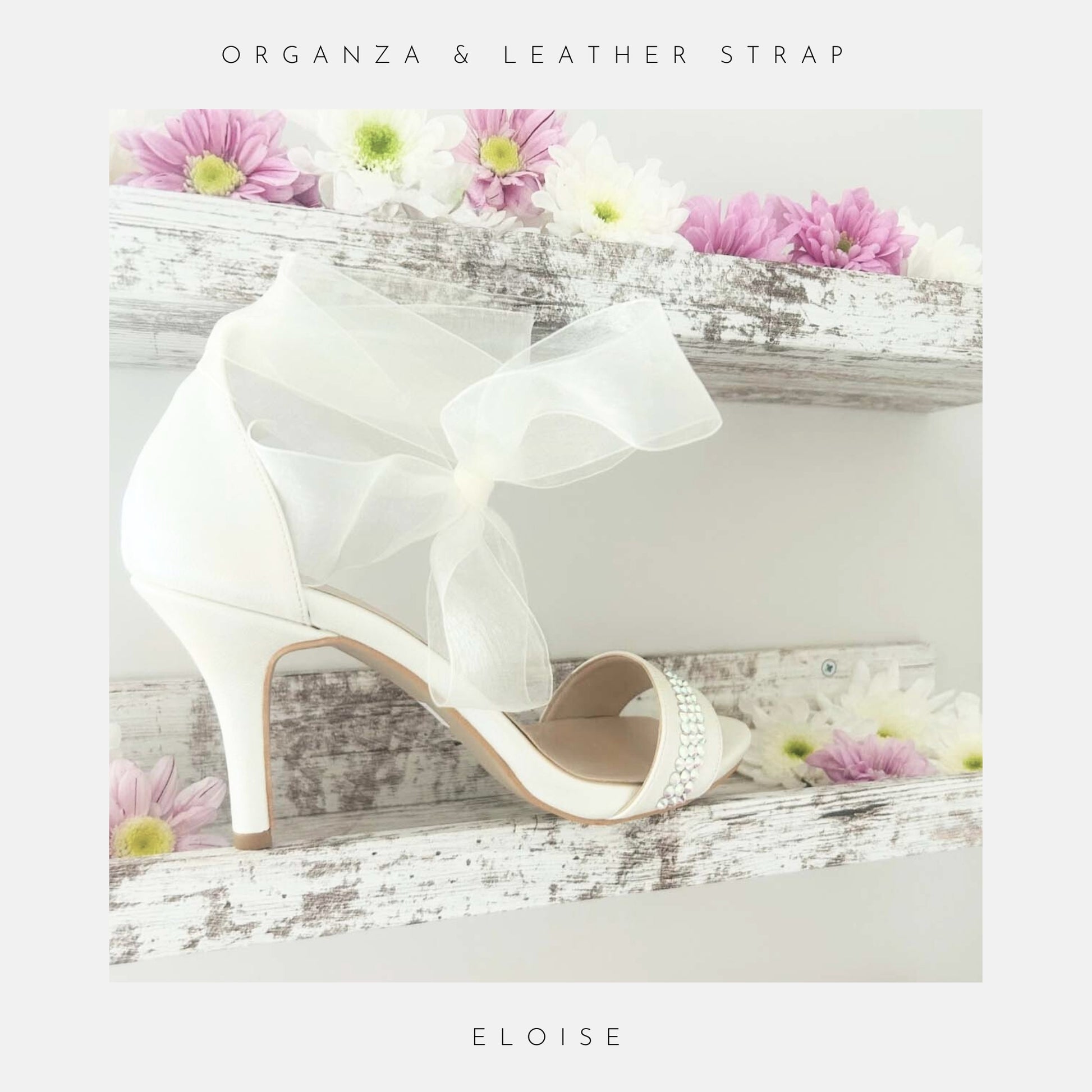 Ivory high-heeled bridal shoes with a bow, placed on a wooden surface with flowers around.