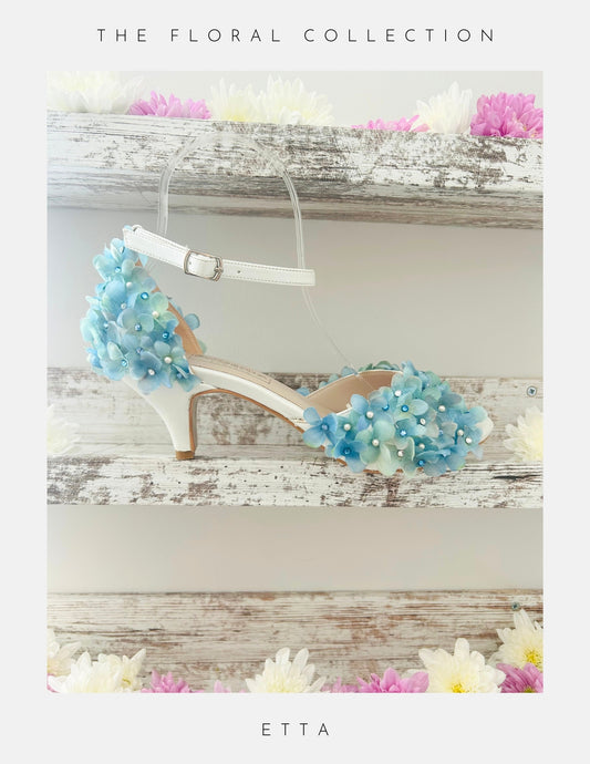 Ivory peep toe bridal sandal with blue floral decorations on a wooden surface with flowers around.