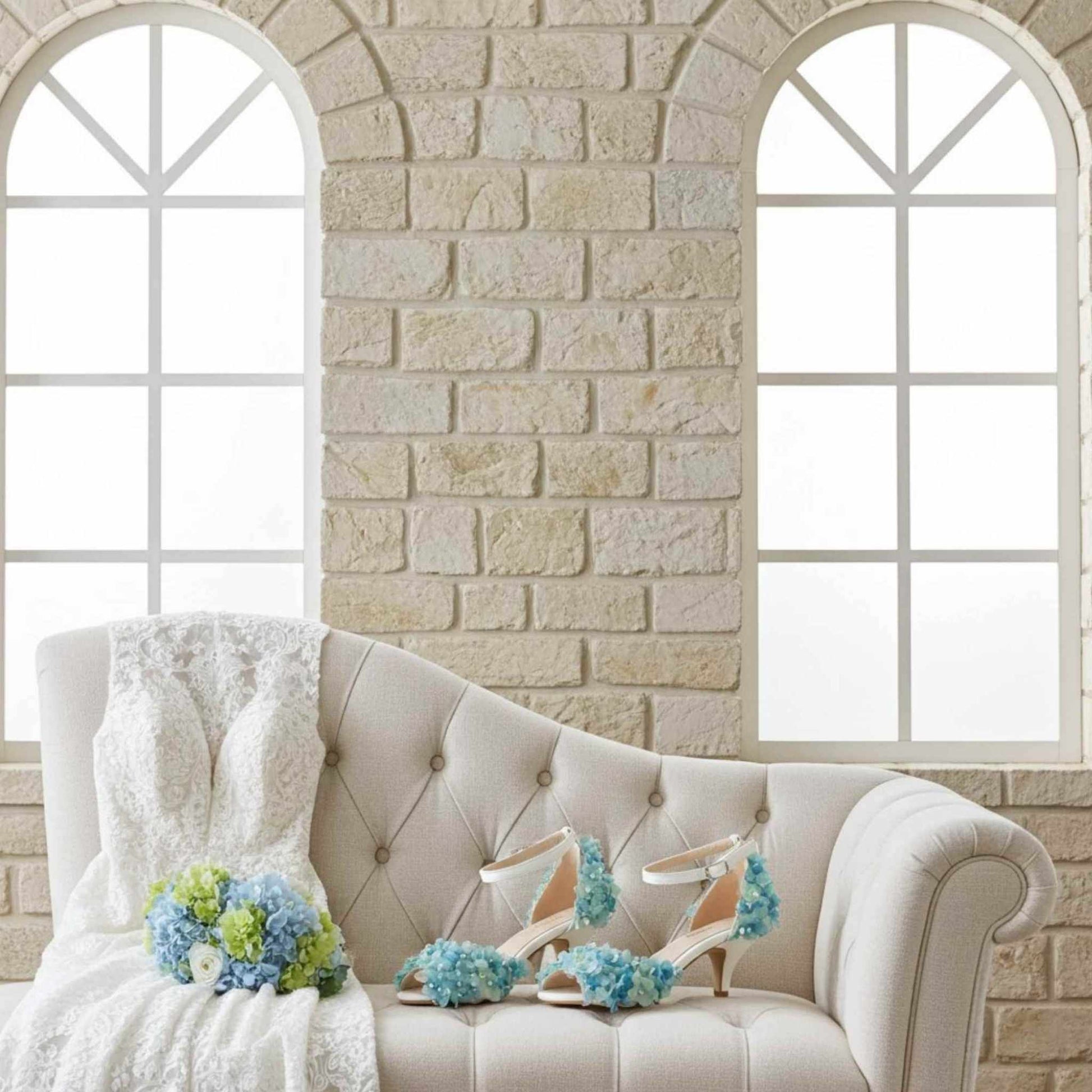 Side view of kitten heel bridal shoes decorated with flowers on a chaise with a wedding bouquet and bridal gown.