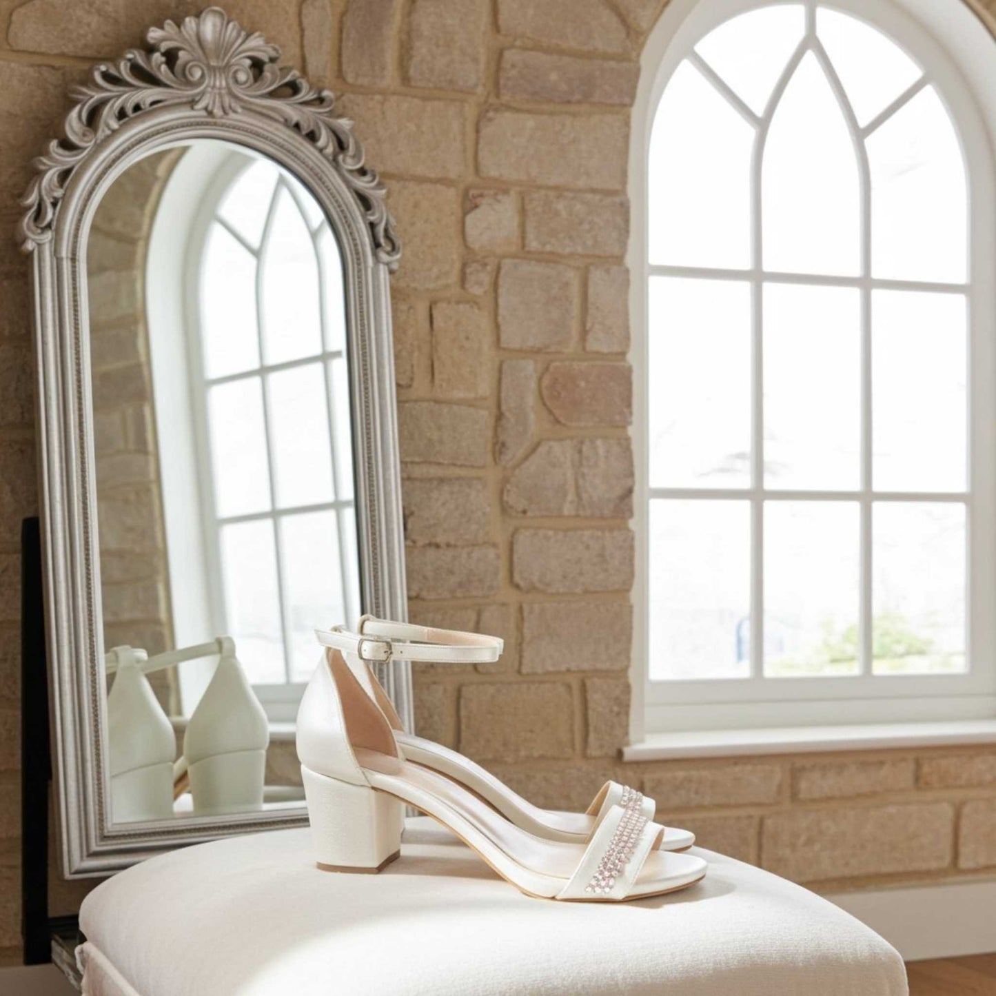 Ivory mid-heeled shoes on a white stool in front of a decorative mirror and stone wall.