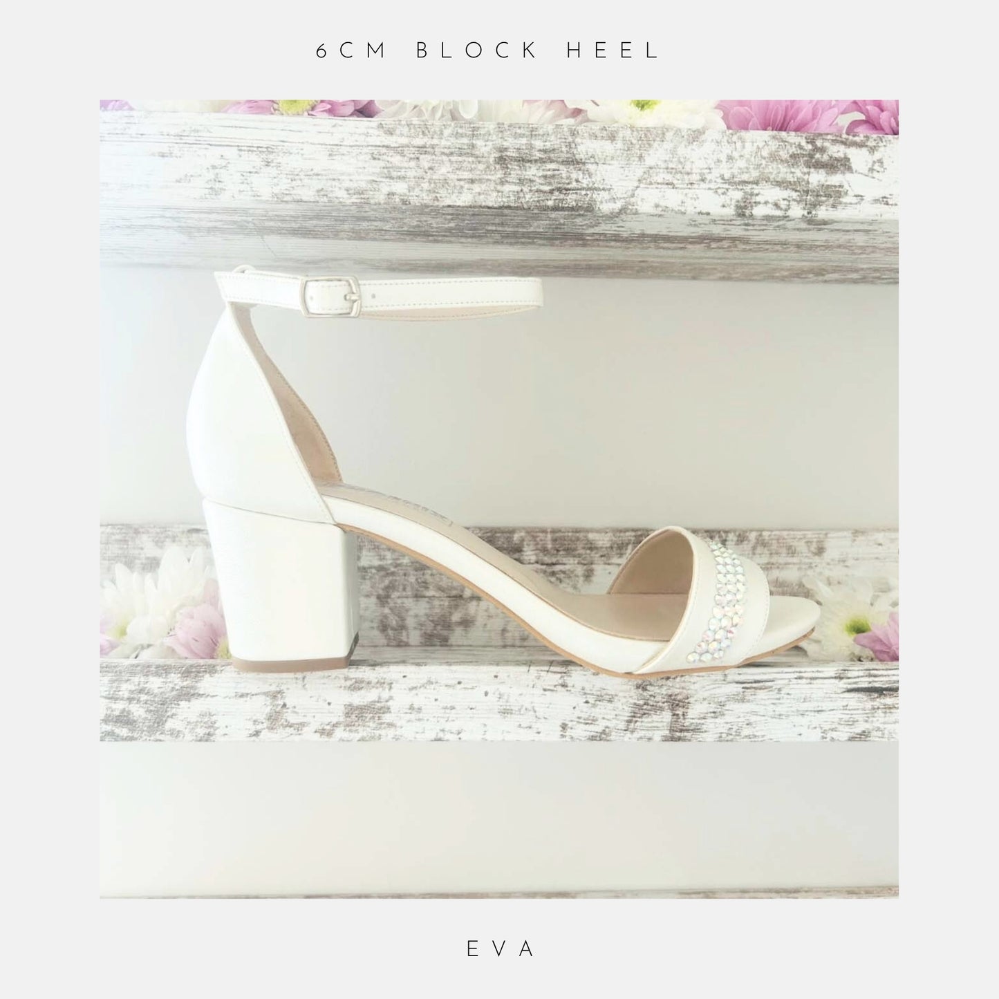 Displayed on a rustic white wooden shelf with pink and white flowers, the Phoenix England Eva Crystal Low Block Heel Bridal Shoes in vegan leather feature a white ankle strap and crystal accents. Text above: 6cm block heel; below: Eva.