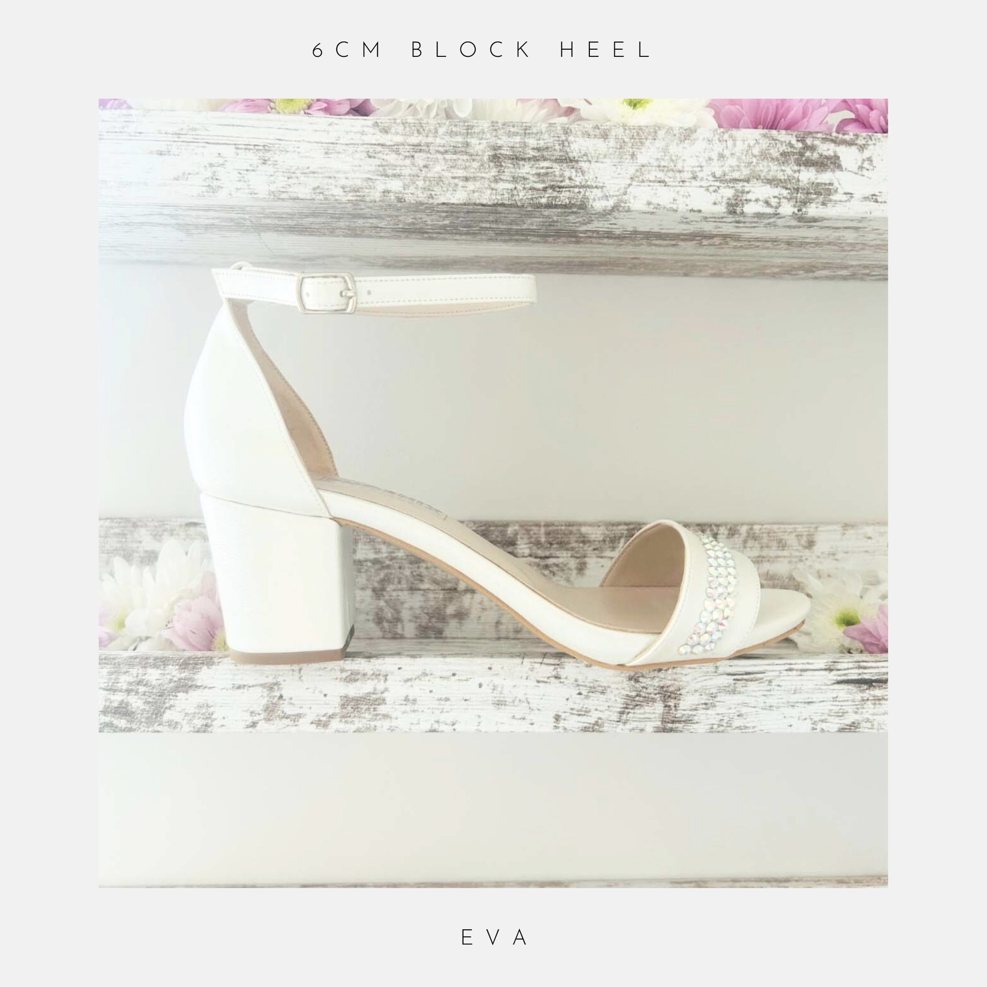 Displayed on a rustic white wooden shelf with pink and white flowers, the Phoenix England Eva Crystal Low Block Heel Bridal Shoes in vegan leather feature a white ankle strap and crystal accents. Text above: 6cm block heel; below: Eva.