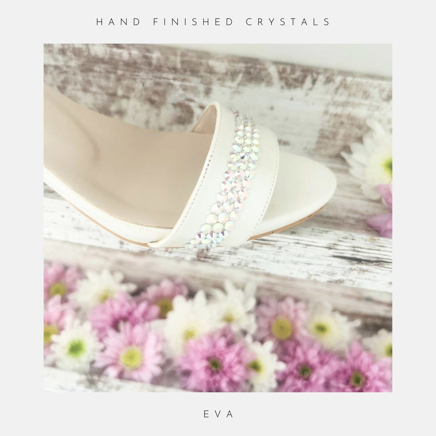 A close-up of the Eva Crystal Low Block Heel Bridal Shoes by Phoenix England, crafted from vegan leather with sparkling crystals on the strap, shown on whitewashed wood with pink and white daisies. Text above: Hand finished with crystals; below: Phoenix England.