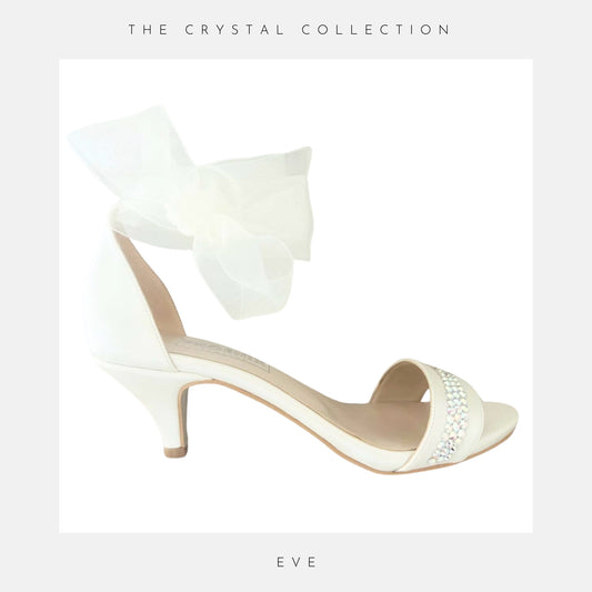 Ivory high-heeled shoe with a large bow and crystal embellishments on a light grey background.