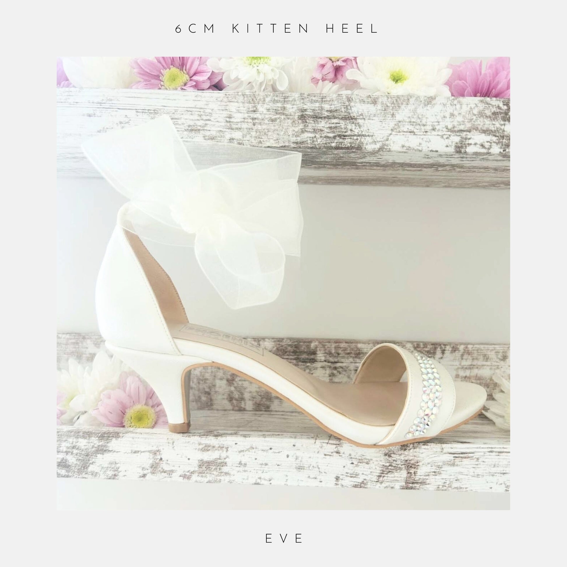 An ivory Phoenix England Eve Organza Kitten Heel Bridal Shoe with a sheer bow and sparkling rhinestones sits on rustic wooden shelves adorned with pink and white daisies. “6cm kitten heel” appears above and “Eve” below.