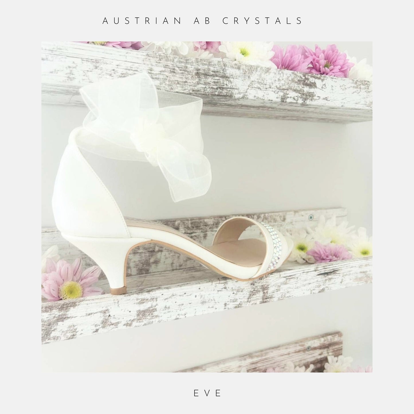 The Phoenix England Eve Organza Kitten Heel Bridal Shoes, featuring a sheer bow and organza strap, are displayed on a rustic white shelf with pink and white flowers. Text above reads Austrian AB crystals and below, Eve.