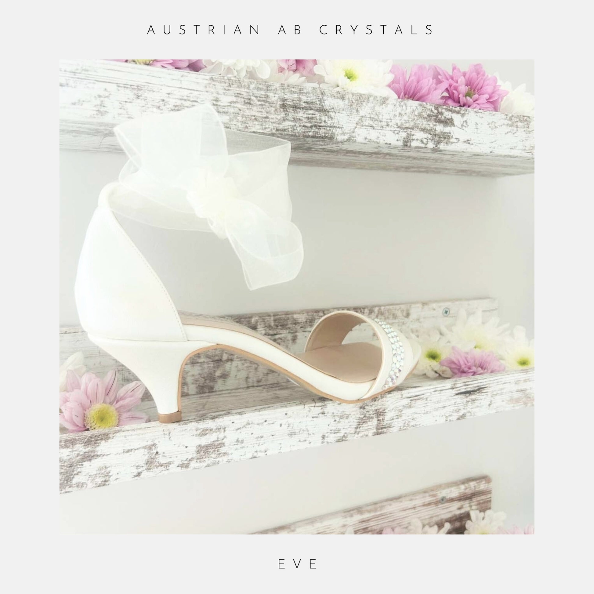 The Phoenix England Eve Organza Kitten Heel Bridal Shoes, featuring a sheer bow and organza strap, are displayed on a rustic white shelf with pink and white flowers. Text above reads Austrian AB crystals and below, Eve.