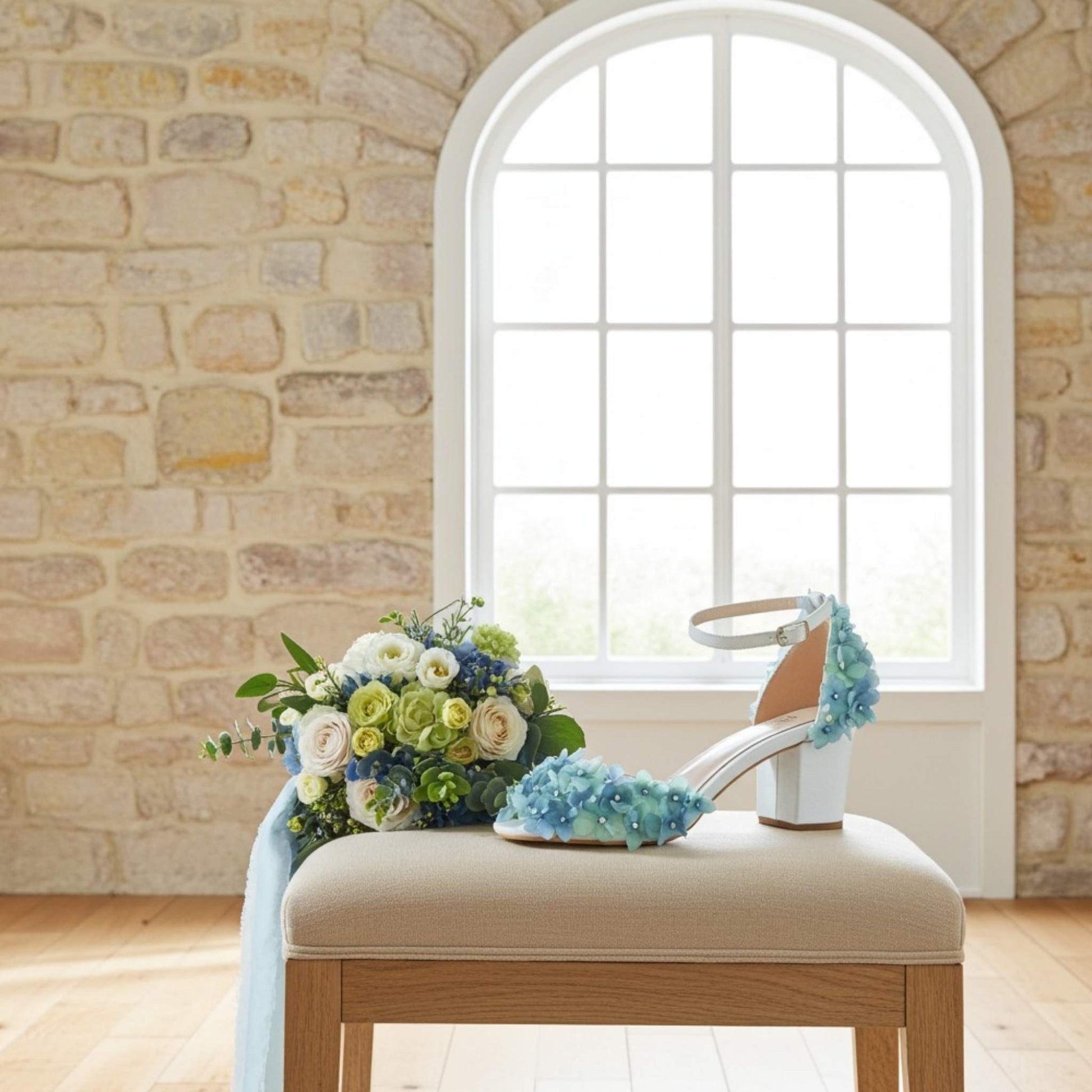Bouquet of flowers and an ivory low block heel sandal with blue floral design on a beige cushion in front of a stone wall and window.