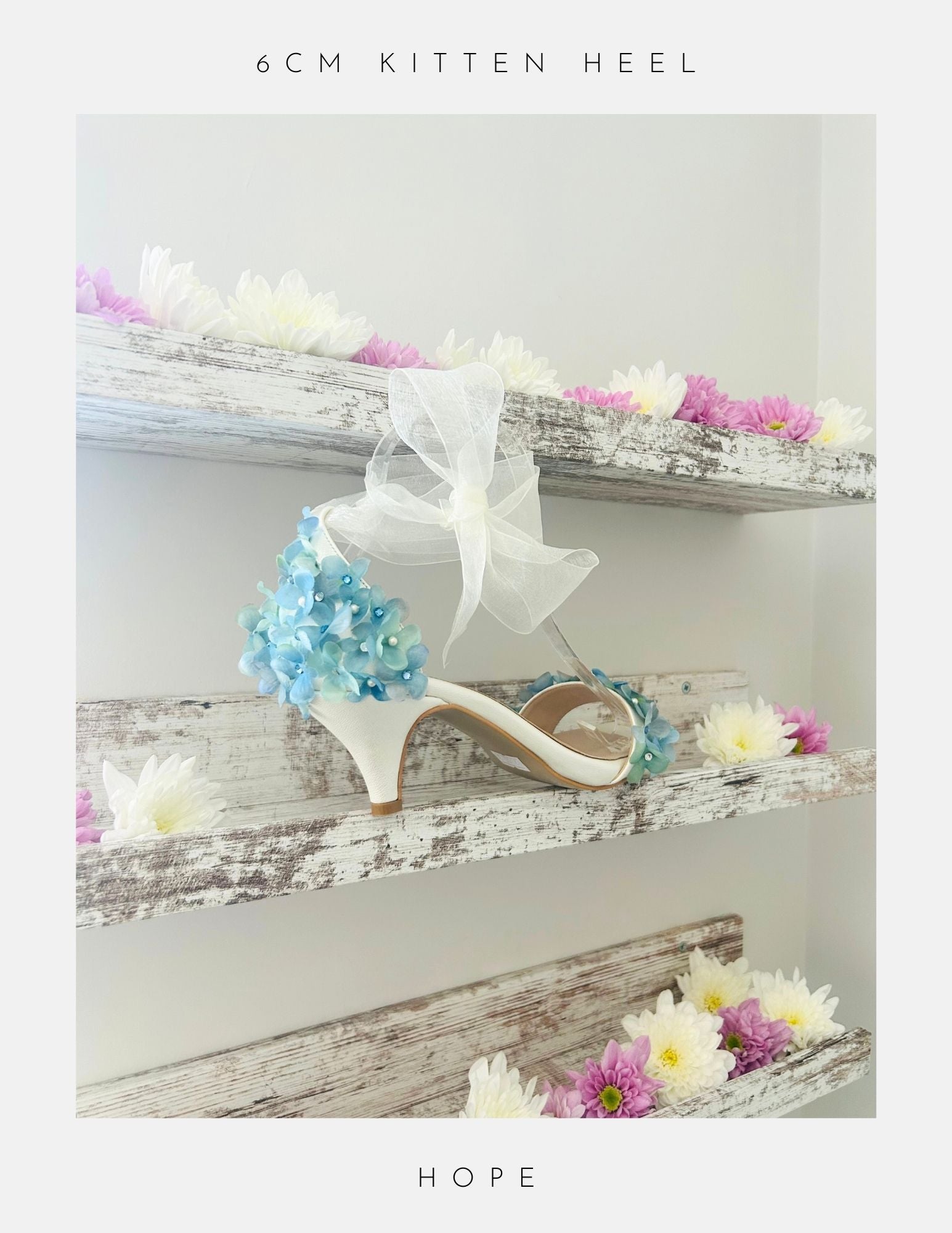 The Hope Organza Floral Kitten Heel Bridal Shoes by Phoenix England feature white kitten heels, blue organza floral accents, and a large white bow, displayed on rustic shelves with pink and white flowers. Text above: 6CM KITTEN HEEL. Below: HOPE.