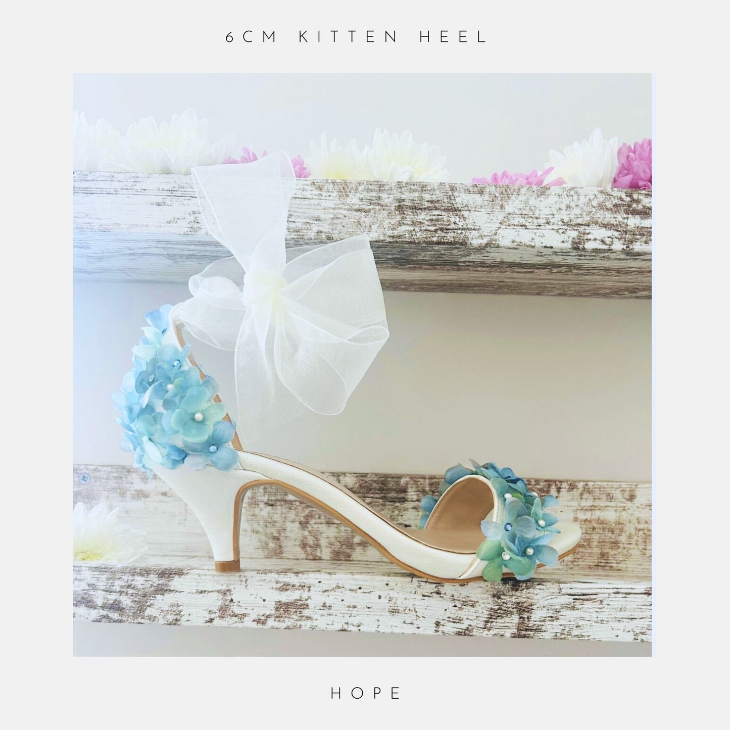 The Hope Organza Floral Kitten Heel Bridal Shoes by Phoenix England, an ivory sandal with blue flowers and a sheer bow rests on a rustic shelf among pastel blooms. Text: 6cm kitten heel and Hope. Perfect for brides.