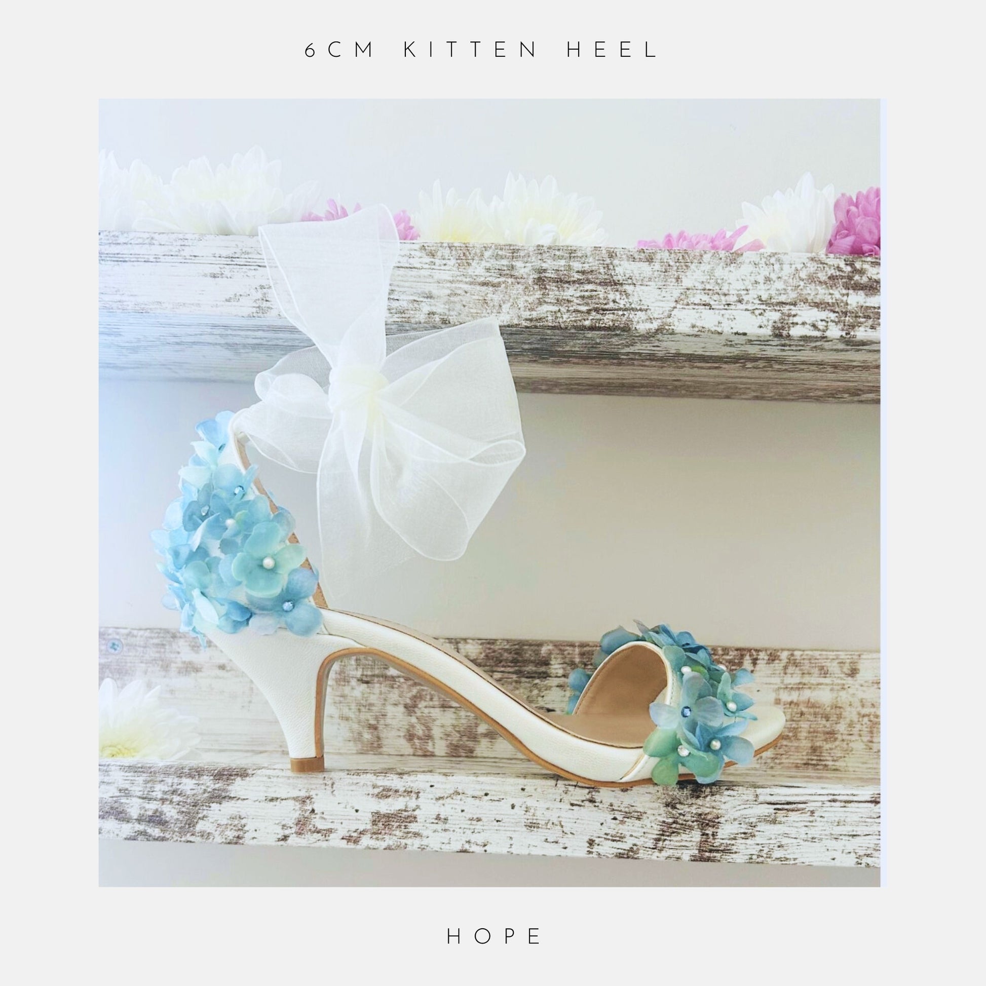 The Hope Organza Floral Kitten Heel Bridal Shoes by Phoenix England, an ivory sandal with blue flowers and a sheer bow rests on a rustic shelf among pastel blooms. Text: 6cm kitten heel and Hope. Perfect for brides.