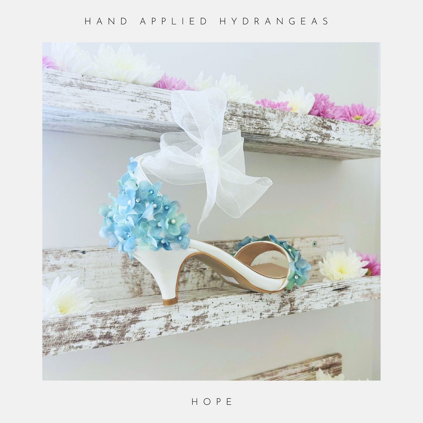 The Hope Organza Floral Kitten Heel Bridal Shoes by Phoenix England feature white kitten heels, blue organza floral accents, and a large ivory bow, displayed on rustic shelves with pink and white flowers. Text above: Hand Applied Hydrangeas. Below: HOPE.