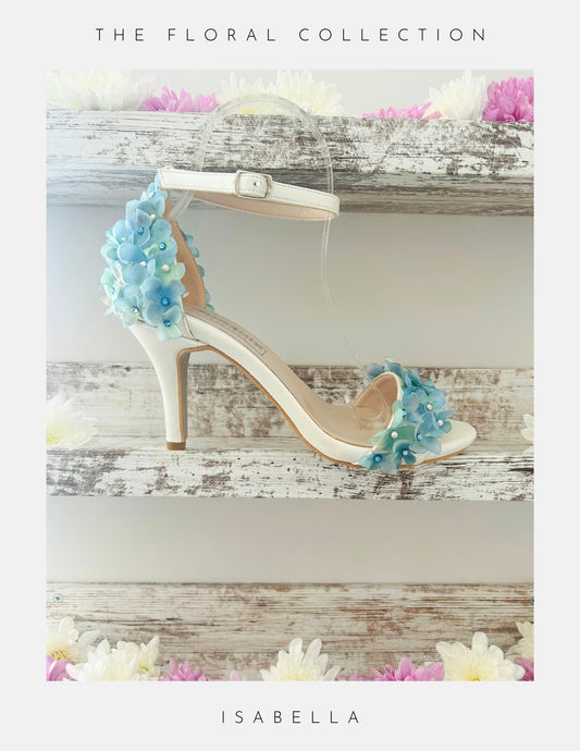 Ivory bridal sandal with floral decorations on a wooden surface, labelled 'The Floral Collection' and 'Isabella'.