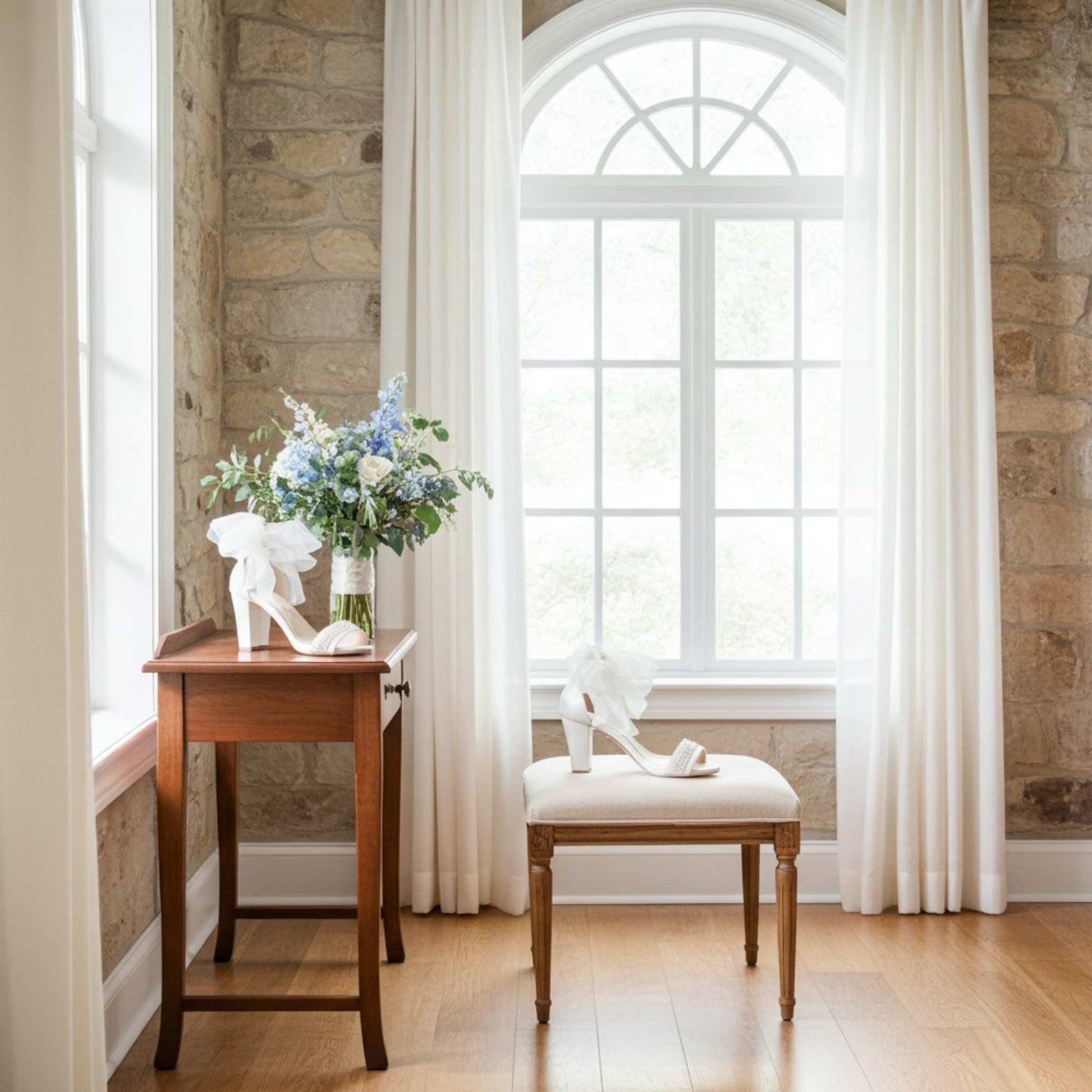 Wooden table with a vase of flowers and ivory organza shoes, next to a cushioned stool in a bright room with stone walls and large windows.