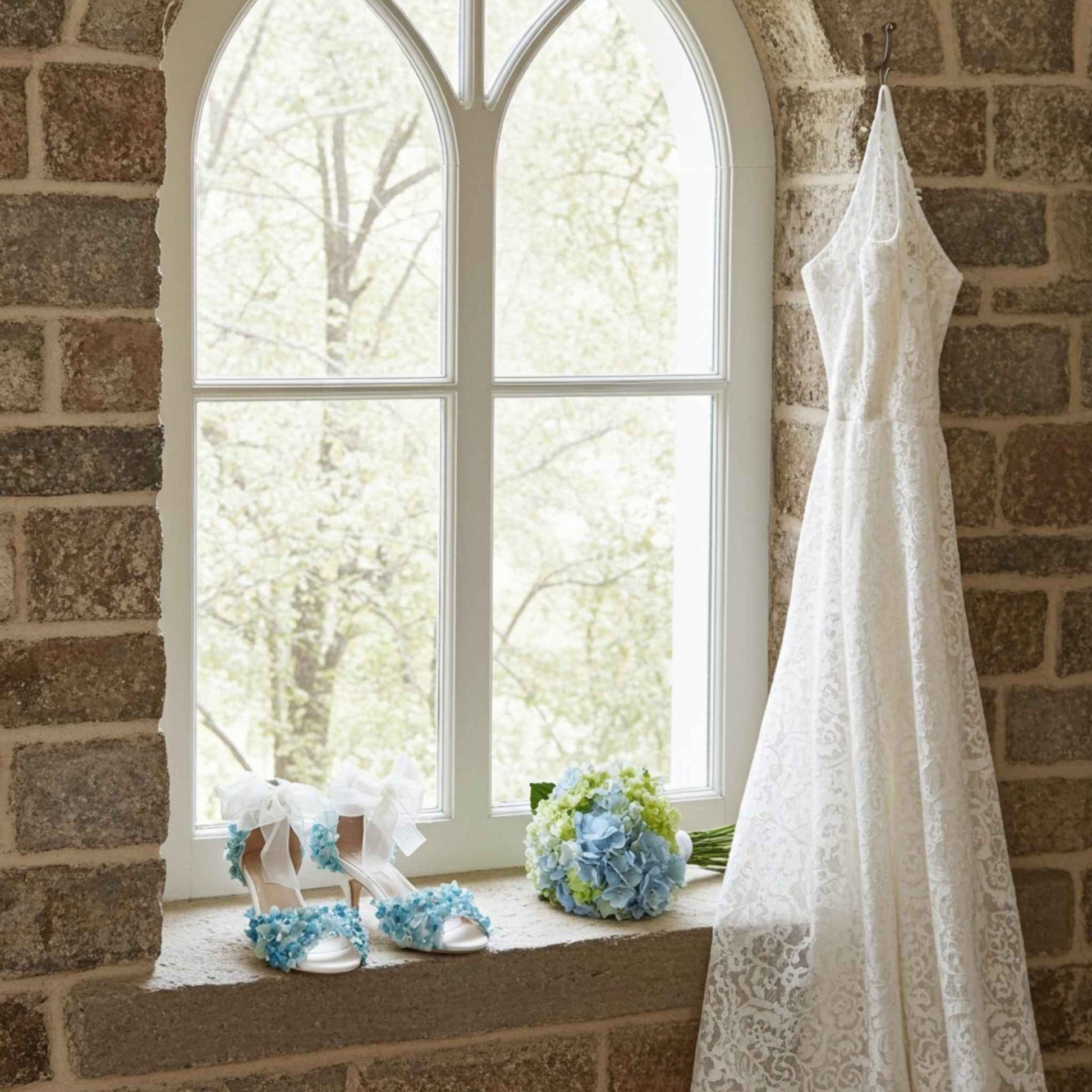 White wedding dress hanging on a brick wall with floral bridal shoes and flowers on a windowsill.