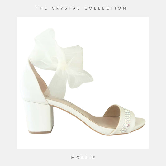 Ivory sandal with bow detail on a light grey background, labelled 'Mollie' under 'The Crystal Collection'.