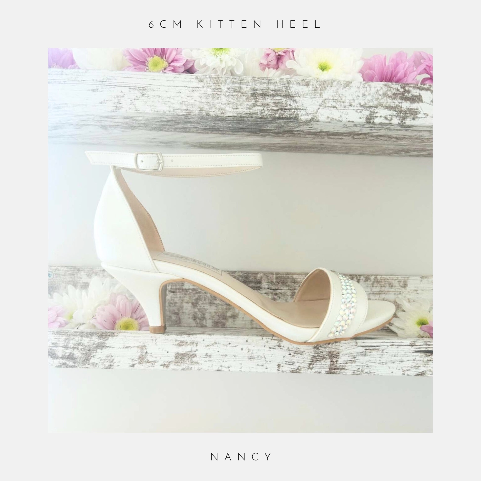 An ivory, open-toe heeled sandal with an ankle strap and crystal accents. The Phoenix England Nancy Crystal Kitten Heel Bridal Shoes, rests on rustic white shelves adorned with pink and white daisies.