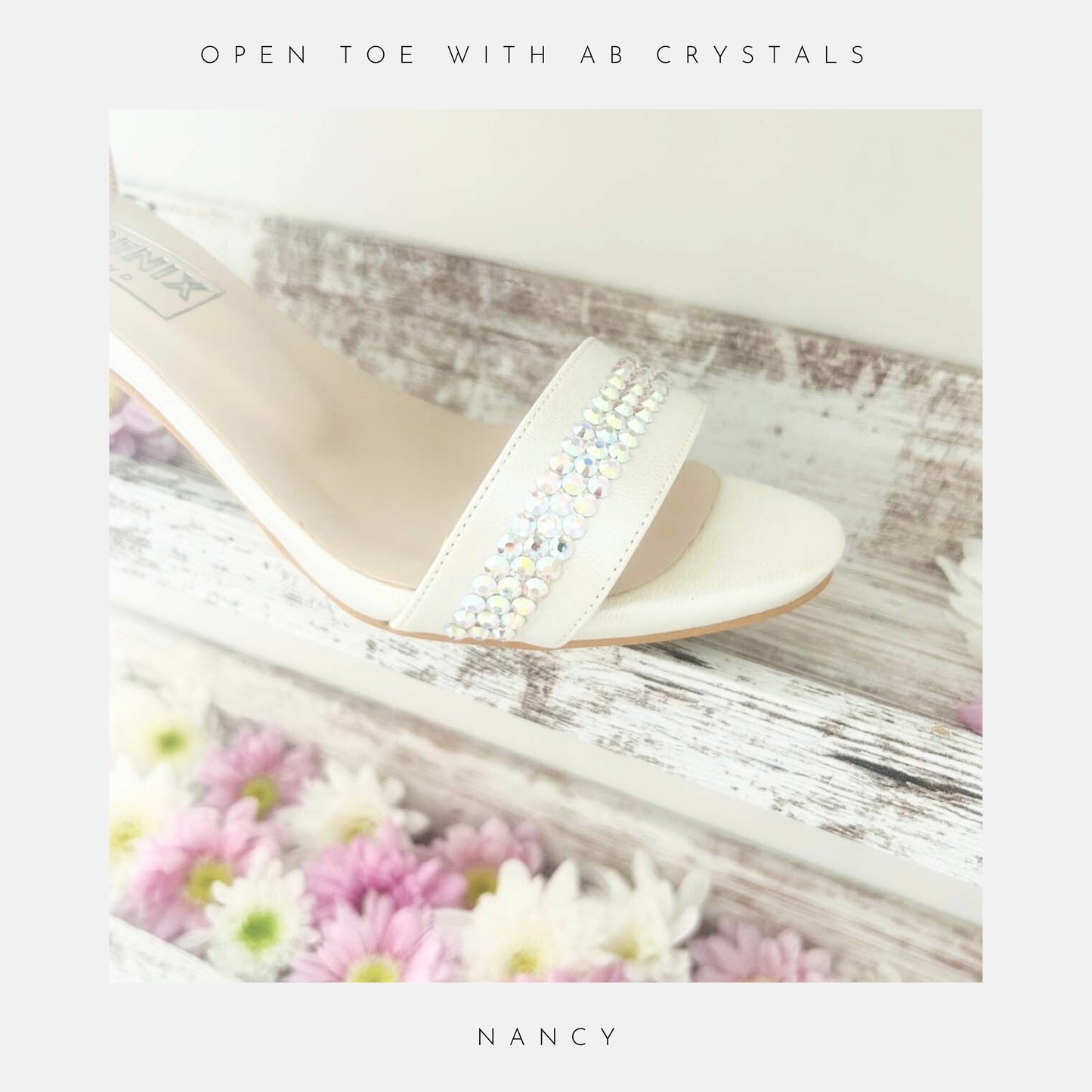 An ivory sandal, the Nancy Crystal Kitten Heel Bridal Shoes by Phoenix England, features pearl vegan leather and a crystal embellished strap. Displayed on a rustic white shelf with pastel daisies. “HAND FINISHED WITH CRYSTALS” above and “NANCY” below. Perfect for weddings.